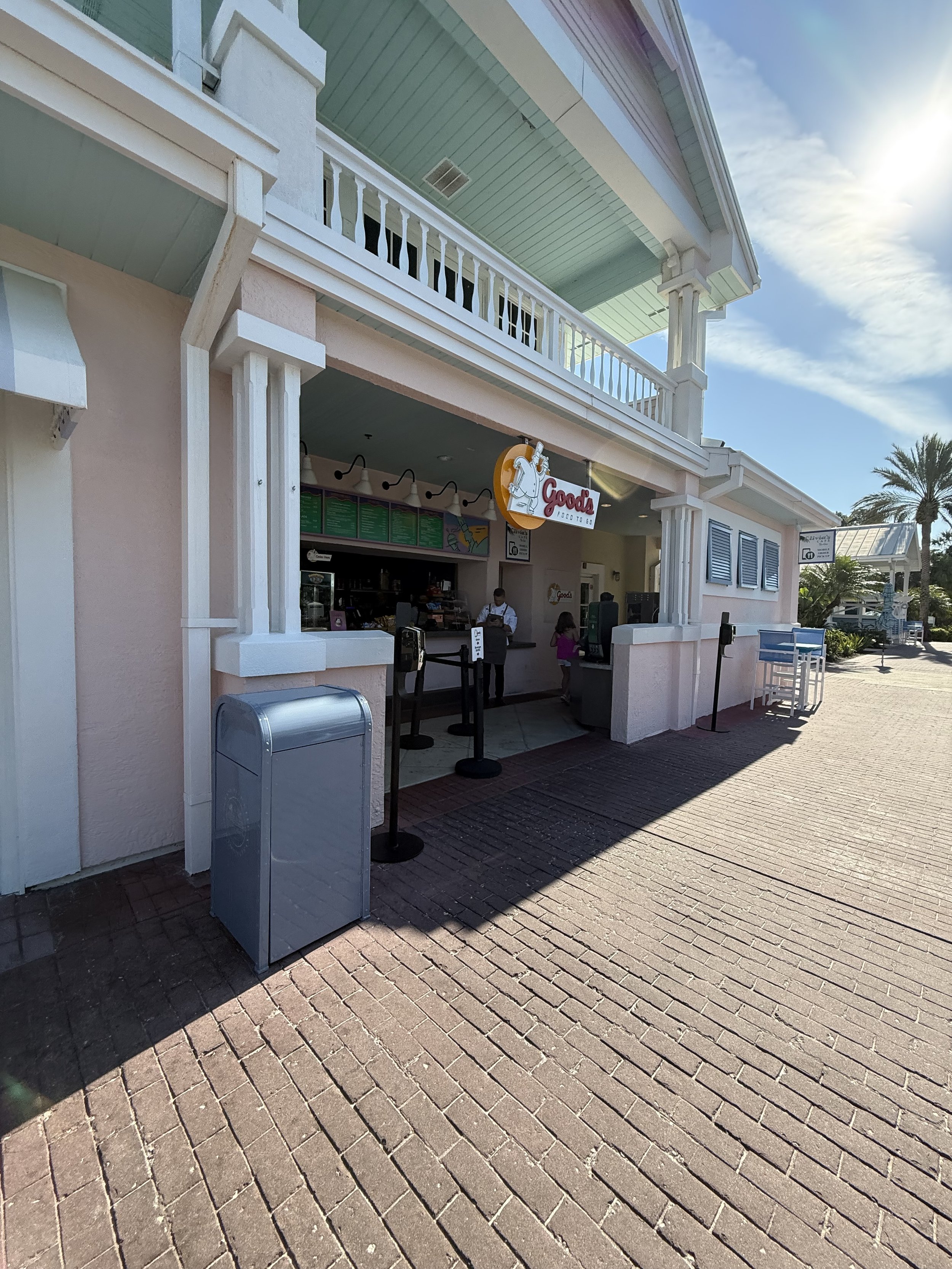 Outside view of a small food stand called 'Goods Food' with a pink building, white accents, and a few people inside. There are palm trees and a bright sunny sky in the background.