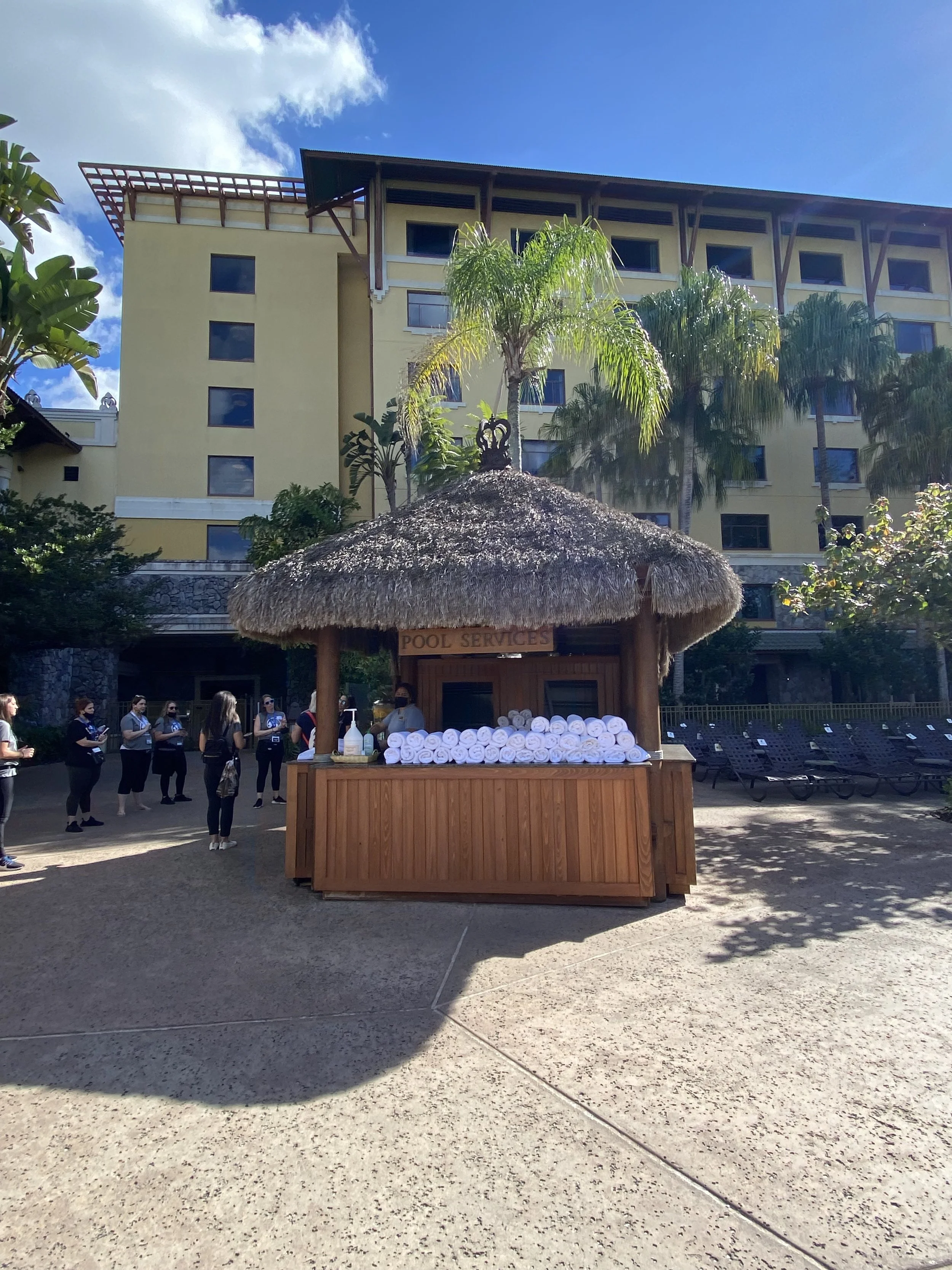 Pool service station with rolled towels and hand sanitizer in front of a hotel building with palm trees.