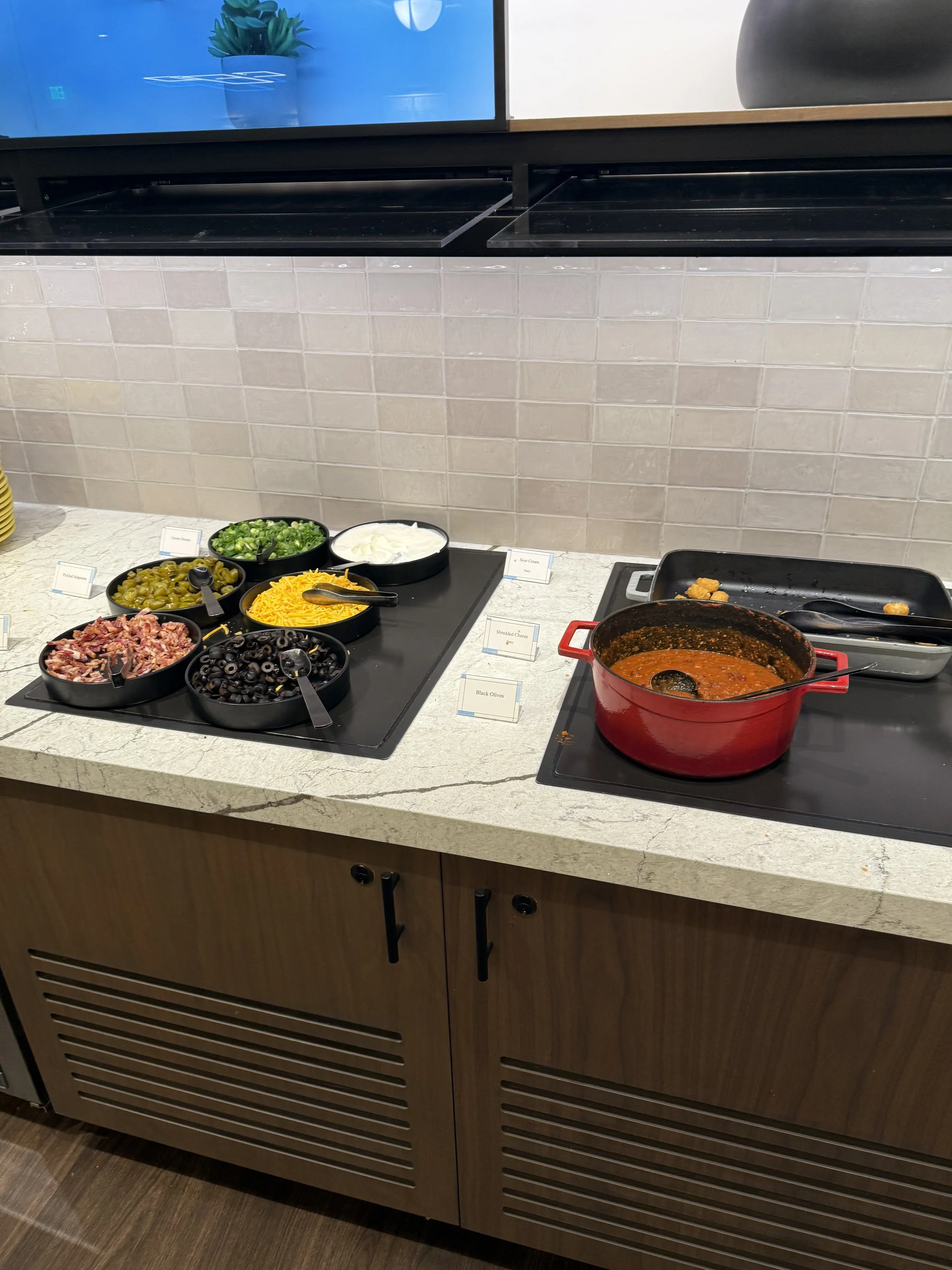 A buffet setup with bowls of toppings including chopped green onions, shredded cheese, sliced black olives, bacon bits, and sour cream, along with a pot of chili and empty baking dishes.