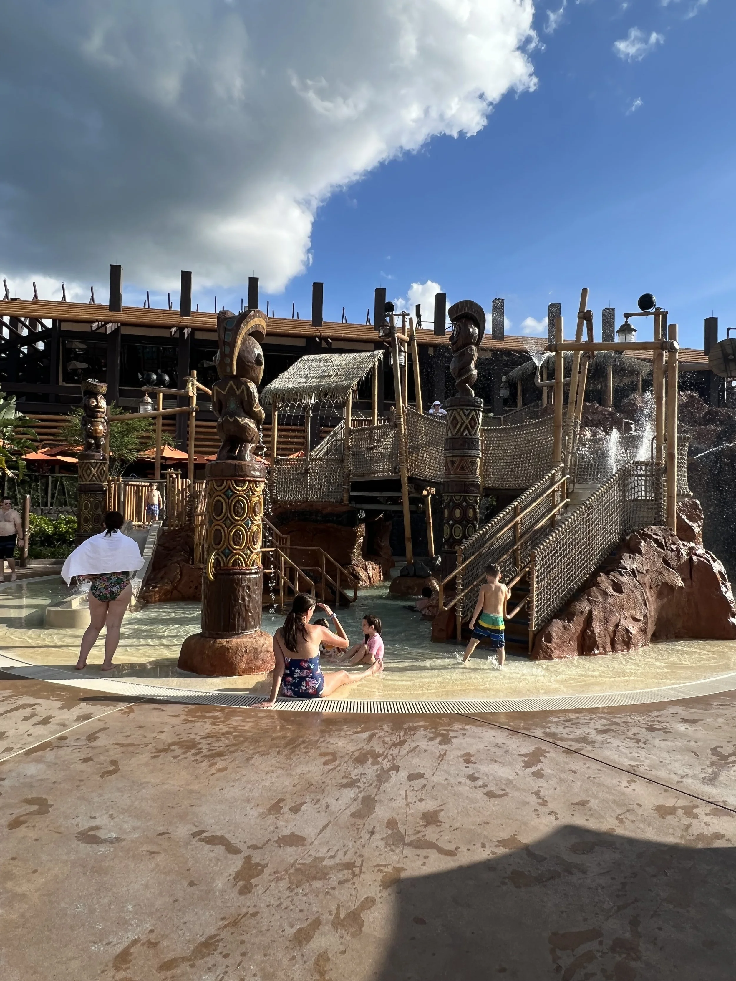 Children and adults playing in a themed water park area with a wooden jungle gym, waterfall features, and Indian tribal carvings, under a partly cloudy sky.