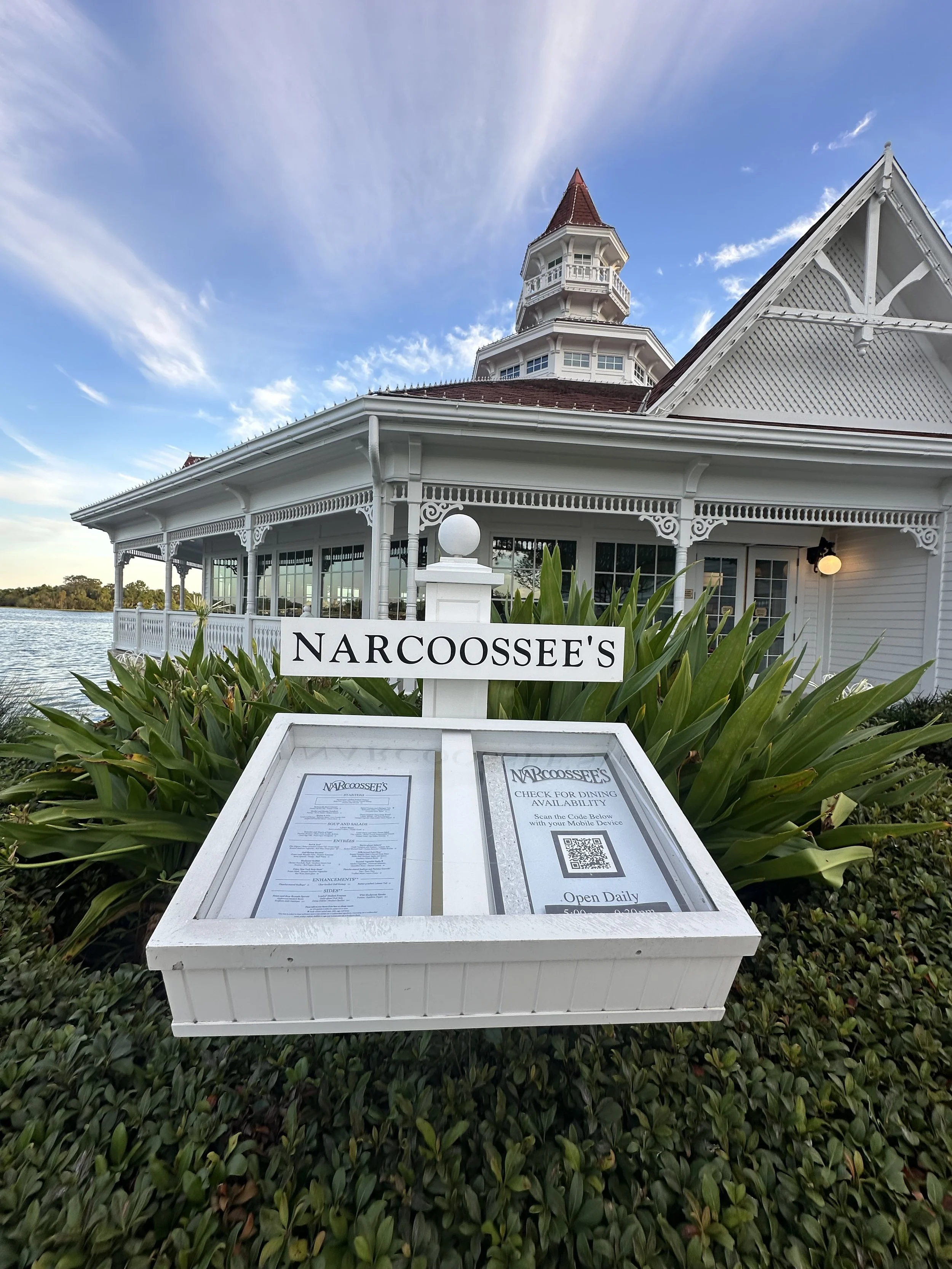 A white historic building with intricate trim and a red-roofed tower, situated by a body of water, with a sign and menu display in front that reads 'NARCOOSSEE'S' and indicates it is open daily for dining.