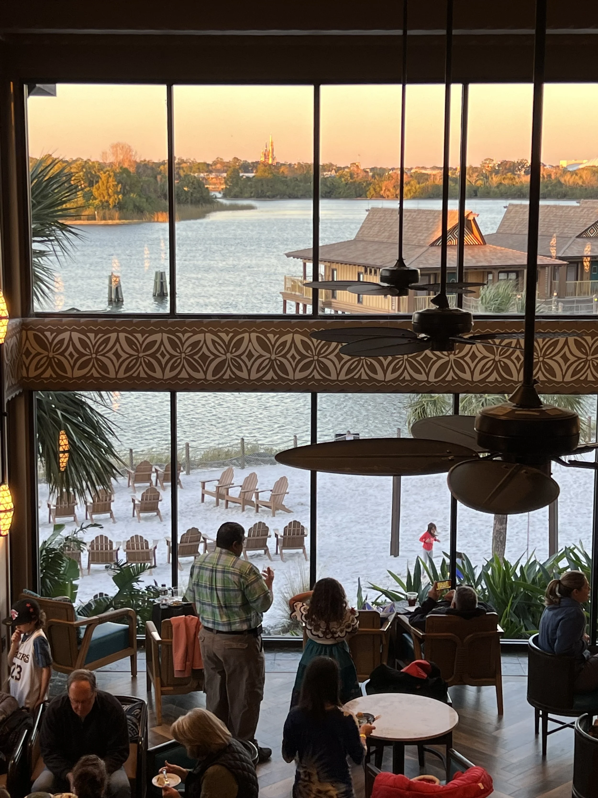 Indoor restaurant with large windows overlooking a lake, with people dining, trees, and houseboats outside, during sunset.