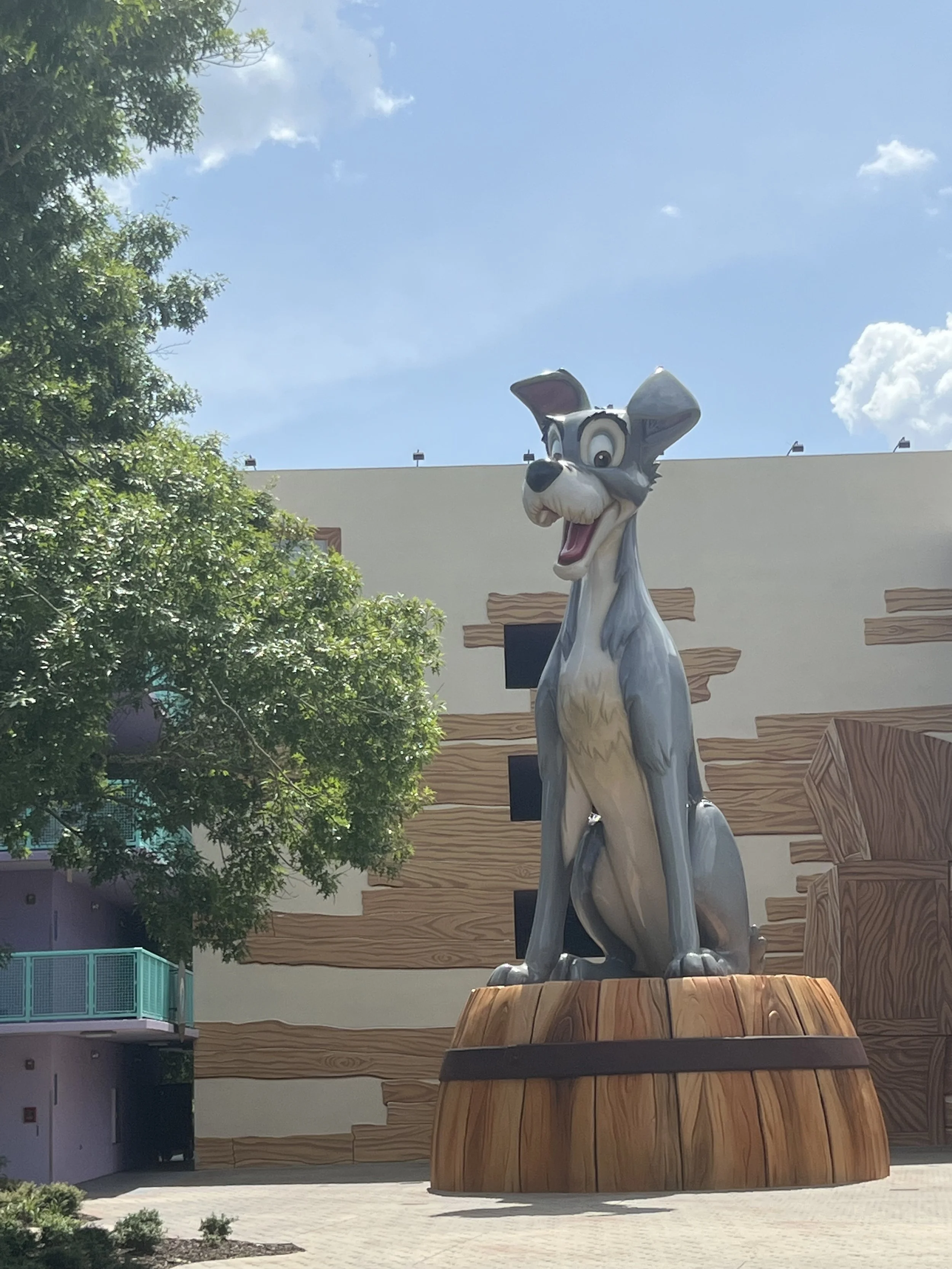 Large cartoon-style statue of a smiling, animated dog, sitting on a wooden platform outdoors under a partly cloudy sky, with trees and a building in the background.