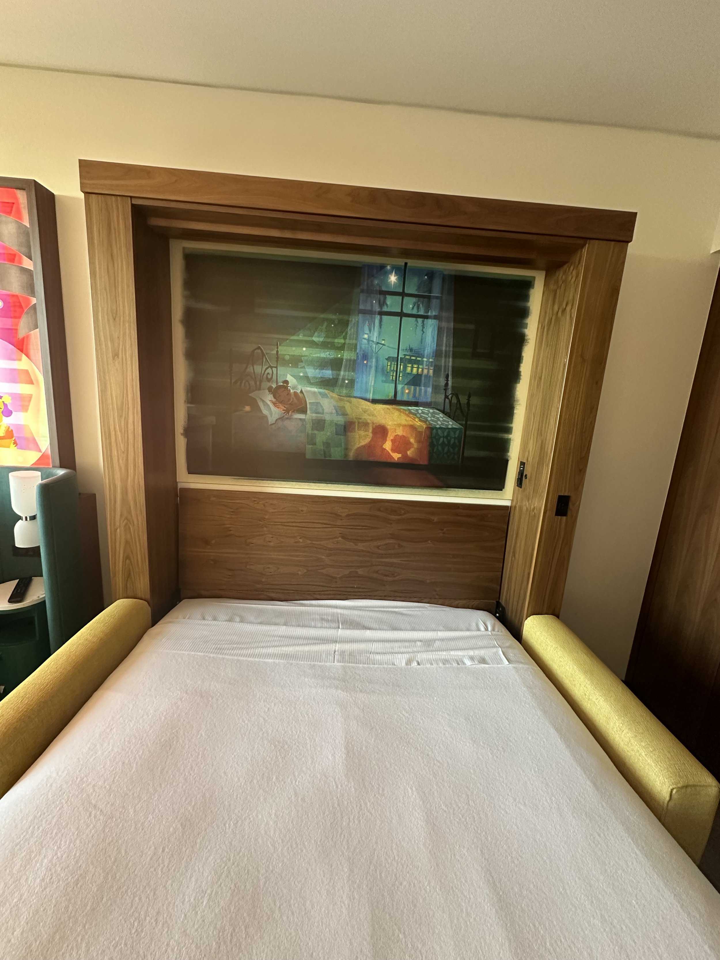 A bed with a headboard and side armrests in a hotel room, featuring a framed picture or screen behind it showing a animated or painted scene of a girl and boy in a room with a window and night sky.
