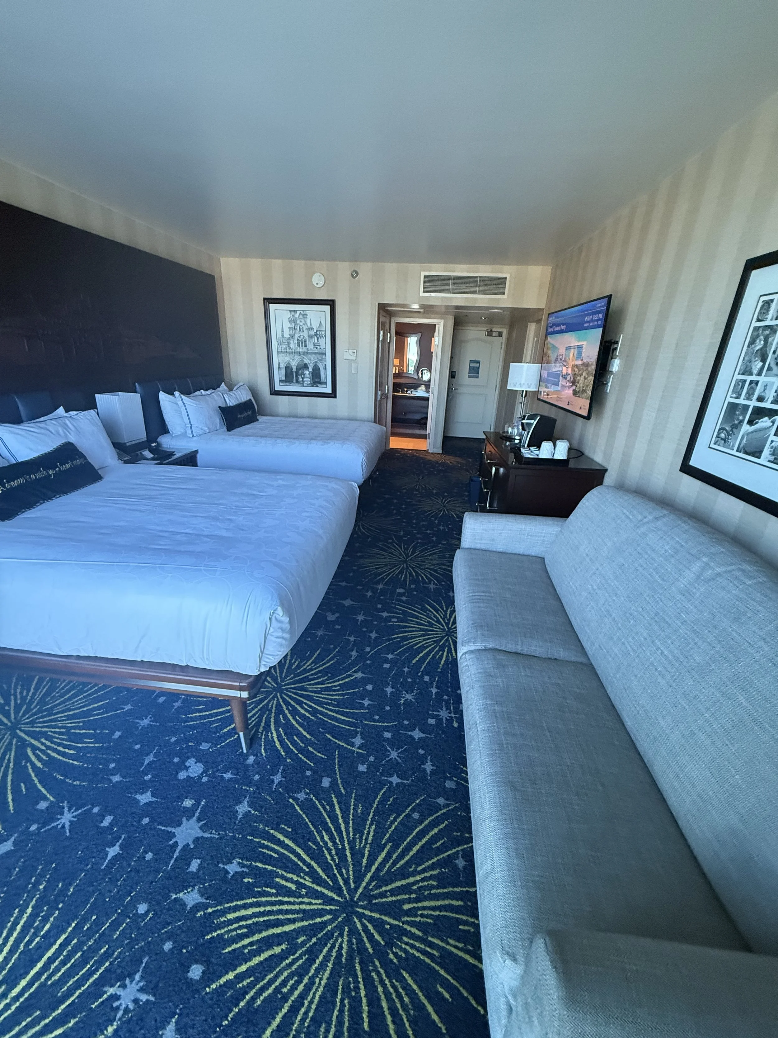 A hotel room with two double beds, a sofa, a flat-screen TV, a lamp, artwork on the walls, and a doorway leading to another room or hallway.