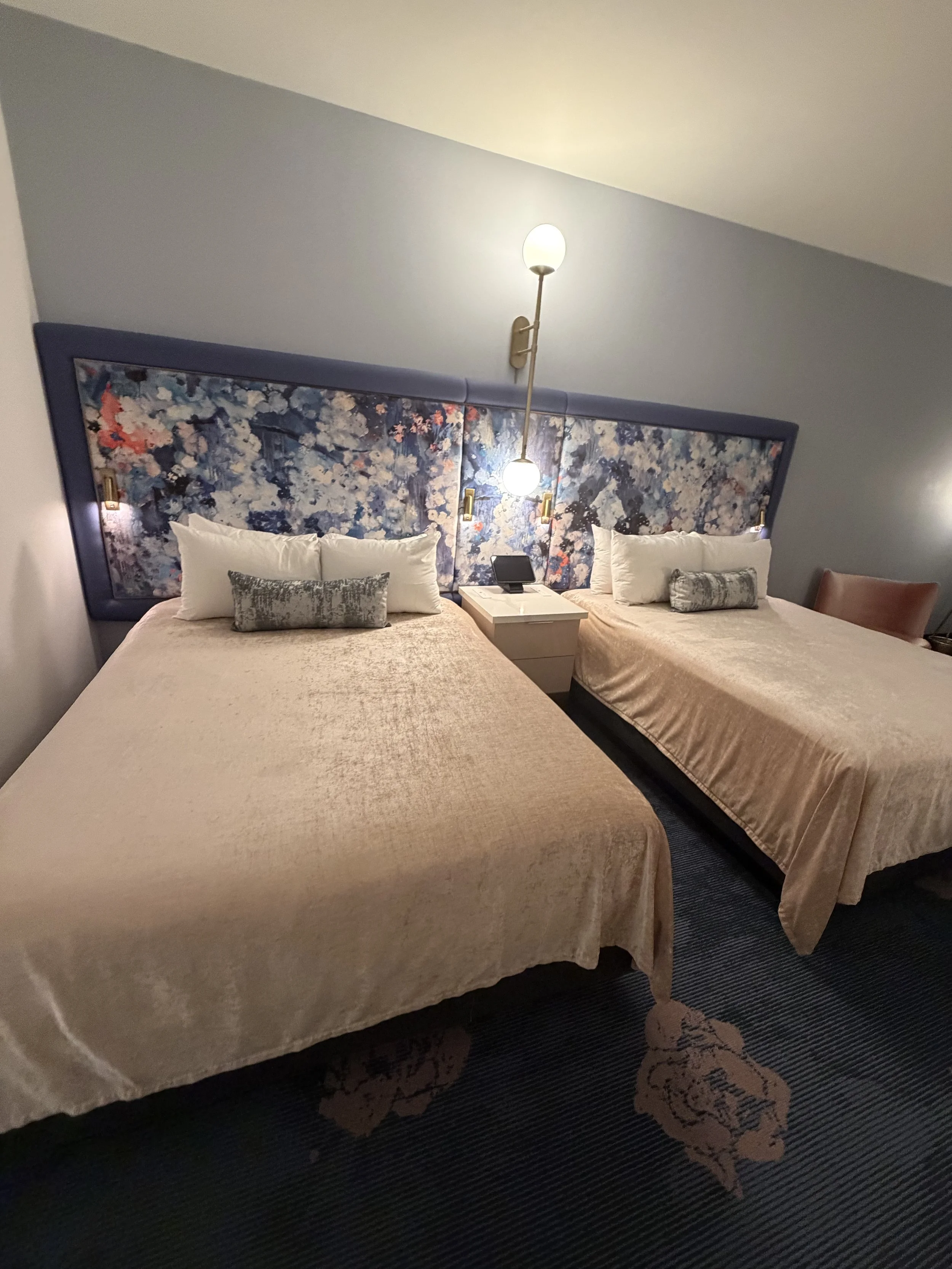 Two beds with beige velvet covers, white pillows, and decorative gray bolster pillows, separated by a nightstand with a black phone, in a hotel room with a floral headboard and blue walls.