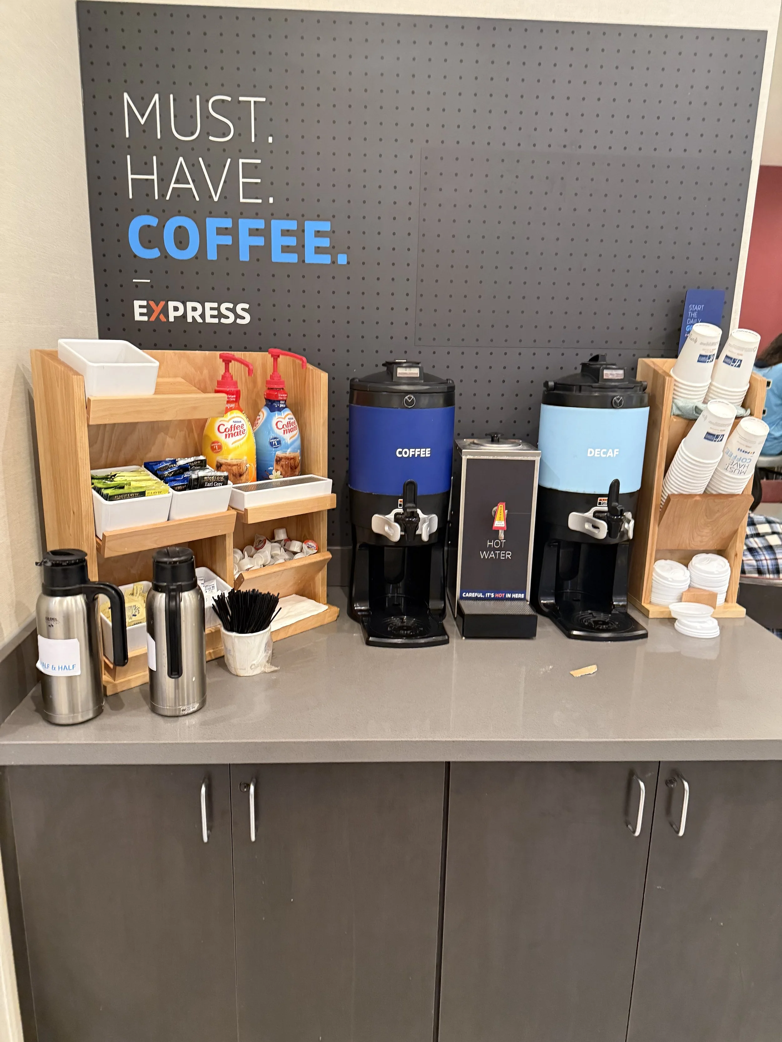 A coffee station with two dispensers labeled 'Coffee' and 'Decaf', a hot water dispenser, and a shelf with cups, stirrers, and miniature bottles of flavored syrups, situated on a gray countertop with gray cabinets below.