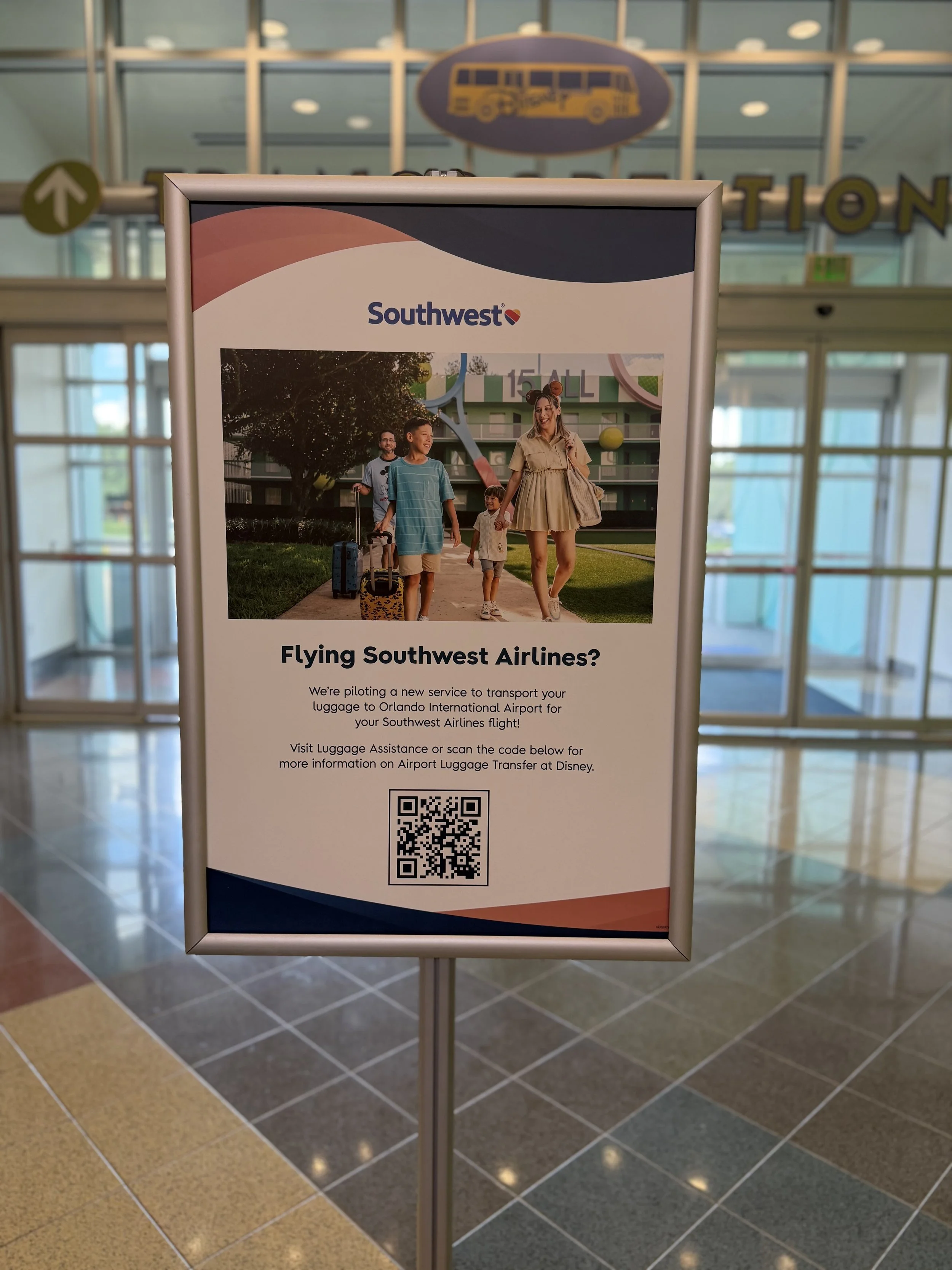 Signboard at the airport announcing Southwest Airlines' new service to Orlando International Airport with a QR code for more information. The sign features a family walking with luggage in front of a Southwest Airlines building.