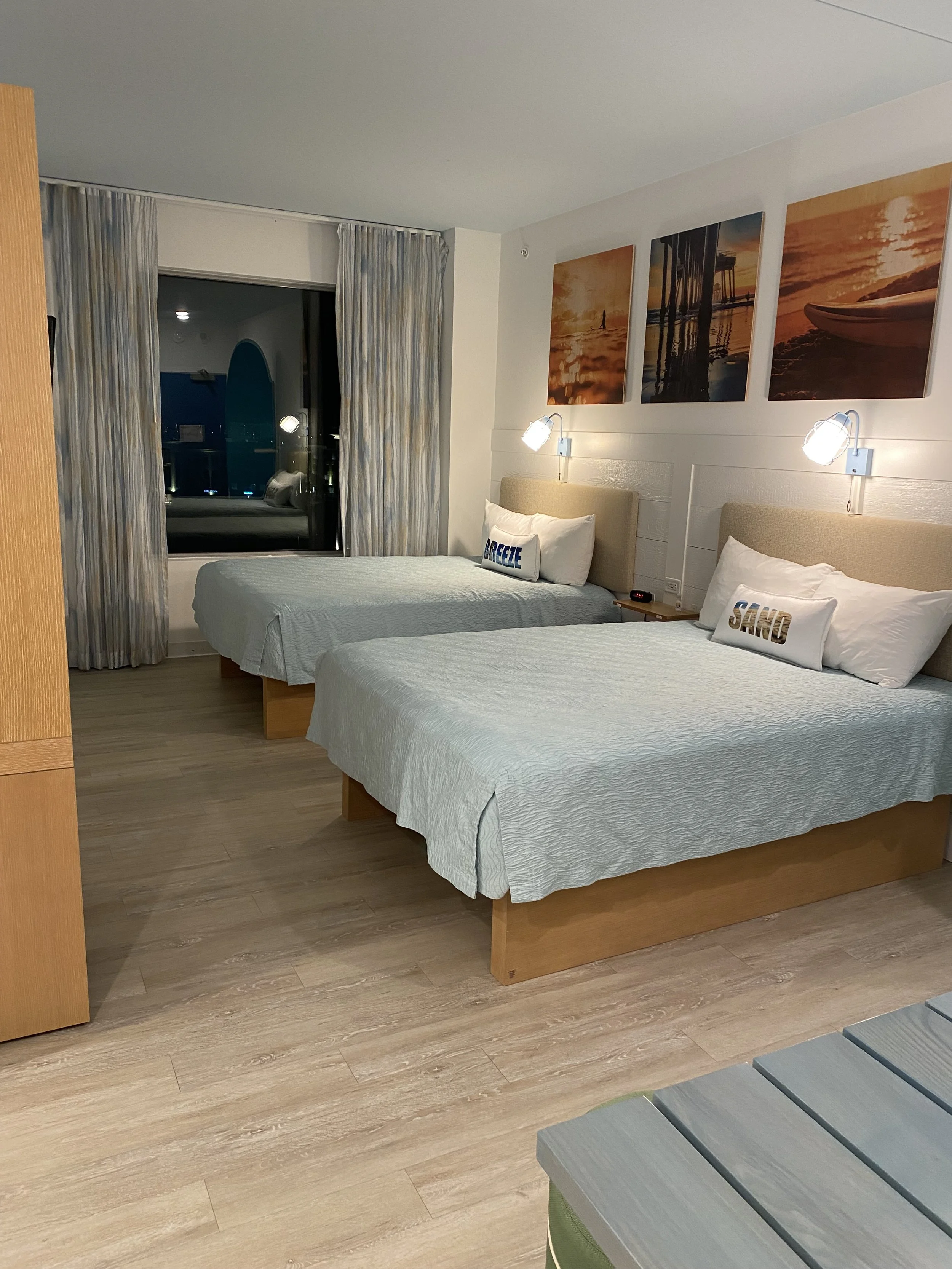 A hotel room with two beds, each with white bedding and pillows that have the words 'BREEZE' and 'SAND' on them. There are two wall-mounted reading lamps above each bed, and three landscape photographs hung above the beds. A window with curtains show