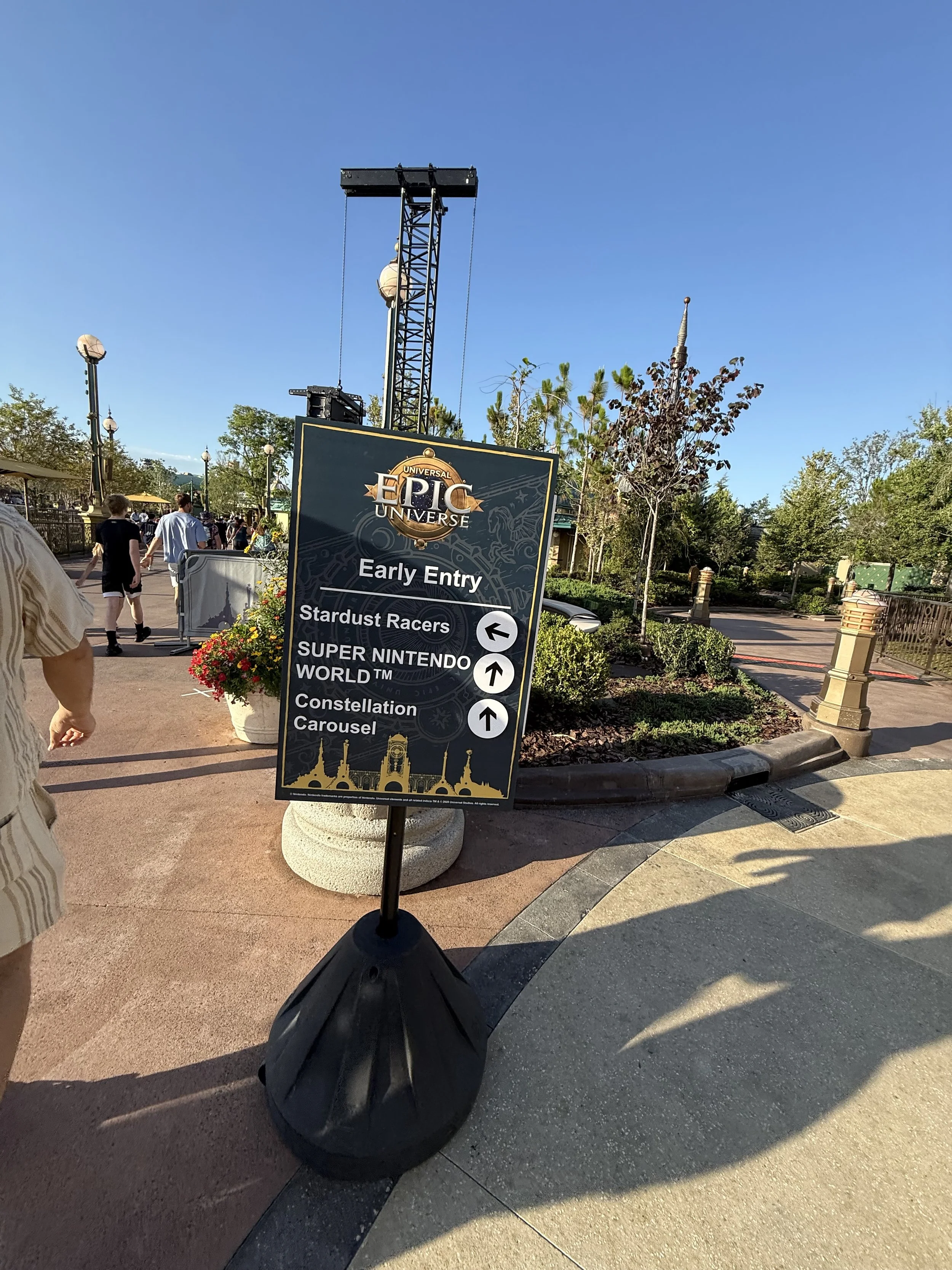 A sign at Disney's EPCOT theme park showing directions for early entry attractions including Stardust Racers, Super Nintendo World, and Constellation Carousel against a backdrop of trees, blue sky, and park visitors.