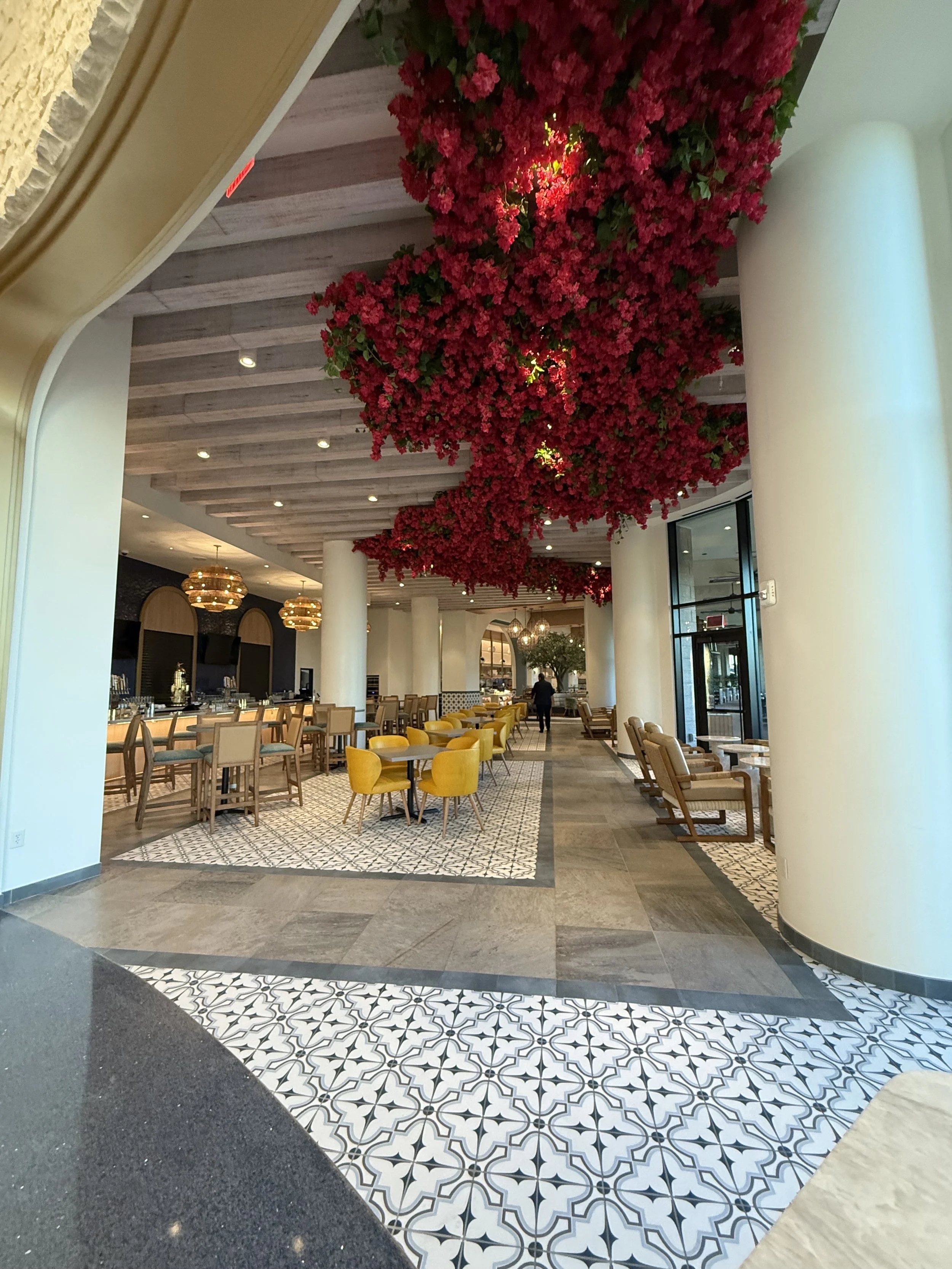 Indoor hotel lobby with a large floral installation of pink flowers hanging from the ceiling, surrounded by seating areas with yellow and beige chairs and patterned floor tiles.