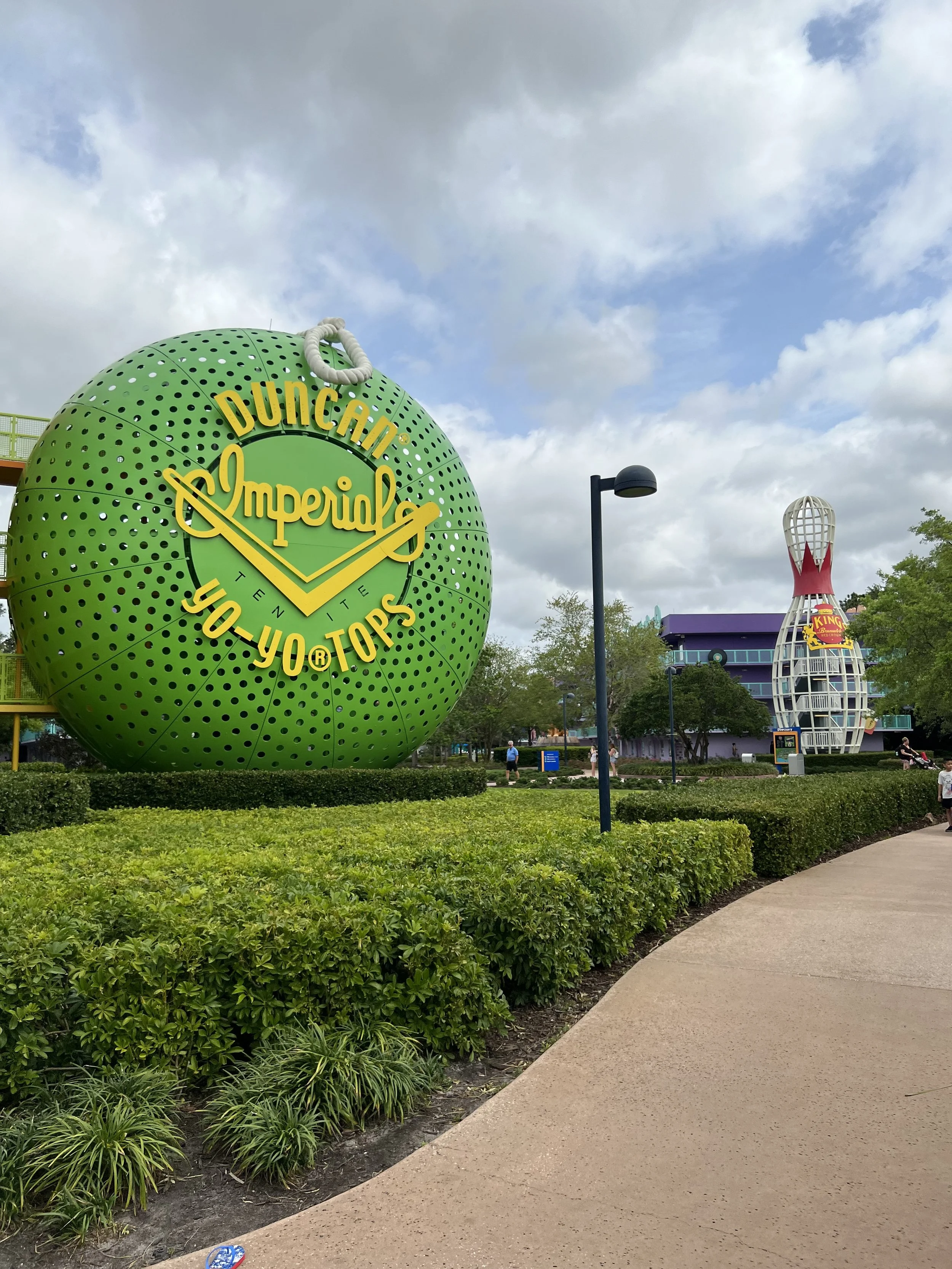 A large green spherical sign with yellow text reads "Duncan Imperial, Tenite, You Topps." In the background, there is a tall, tilted structure and a building, with a sidewalk, trees, and people walking around.