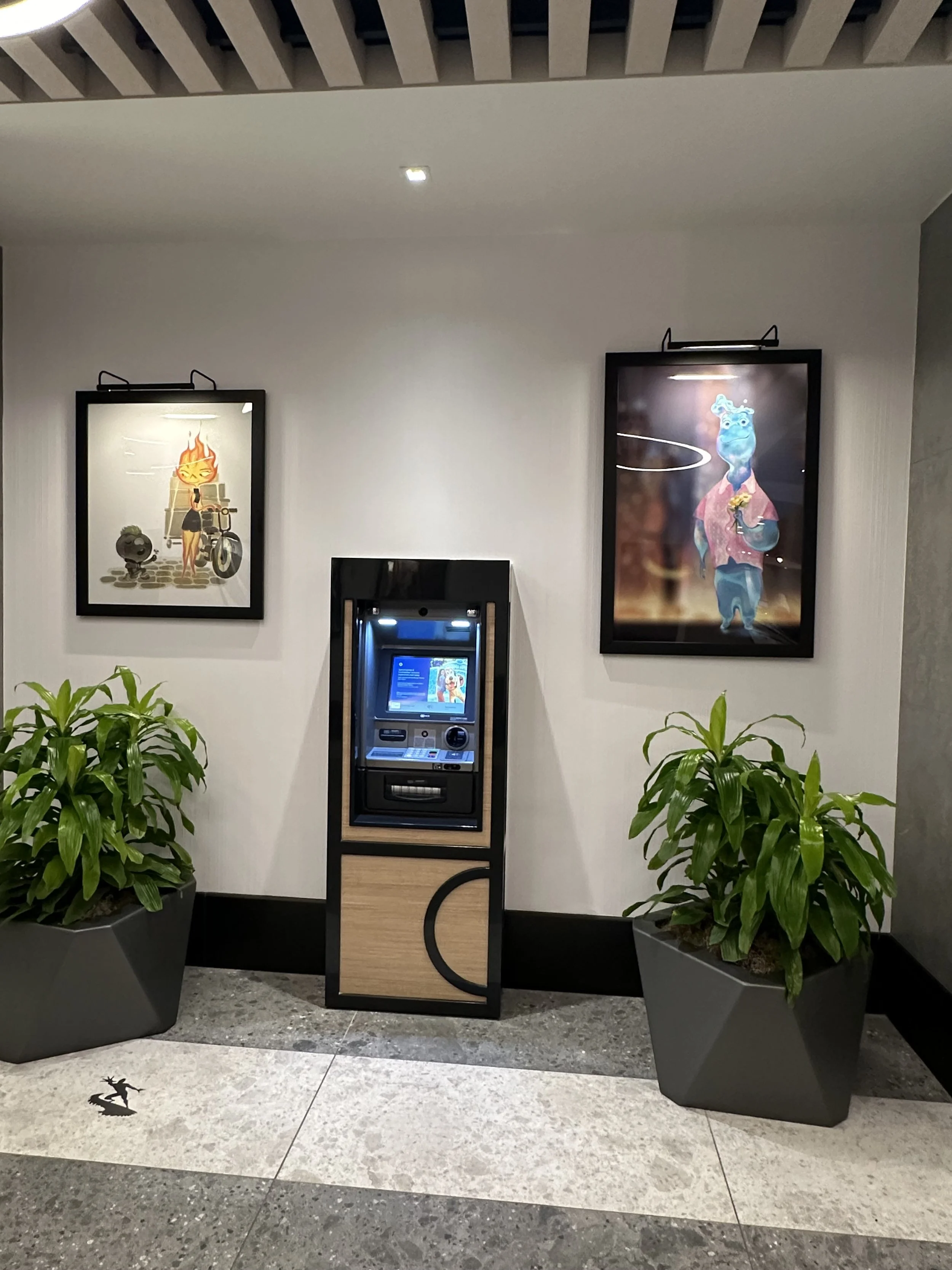 An indoor scene with two framed colorful art posters on a white wall, a modern ATM machine in the center, and two large potted green plants on either side of the ATM. The ceiling has a wooden slat design, and the floor is made of stone tiles.