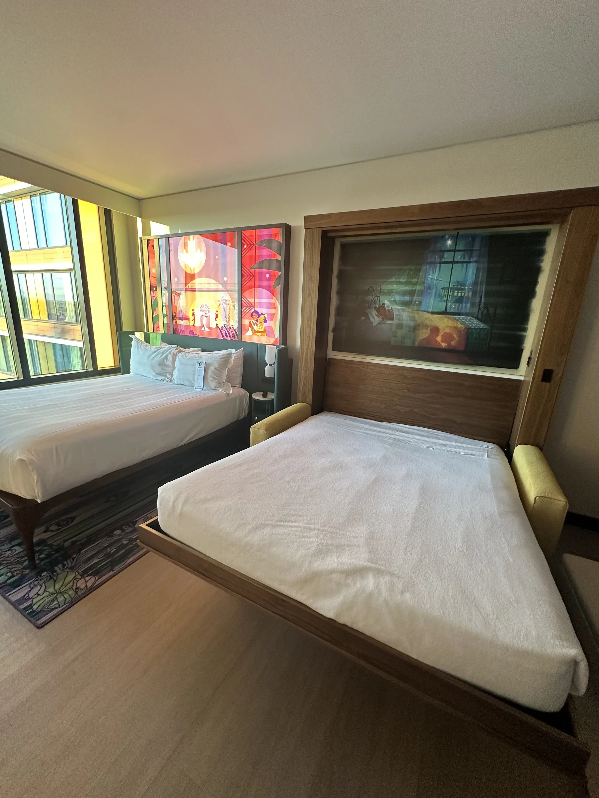 A hotel room with two beds, one with plain white bedding and the other with a patterned headboard, illuminated by colorful stained glass windows.