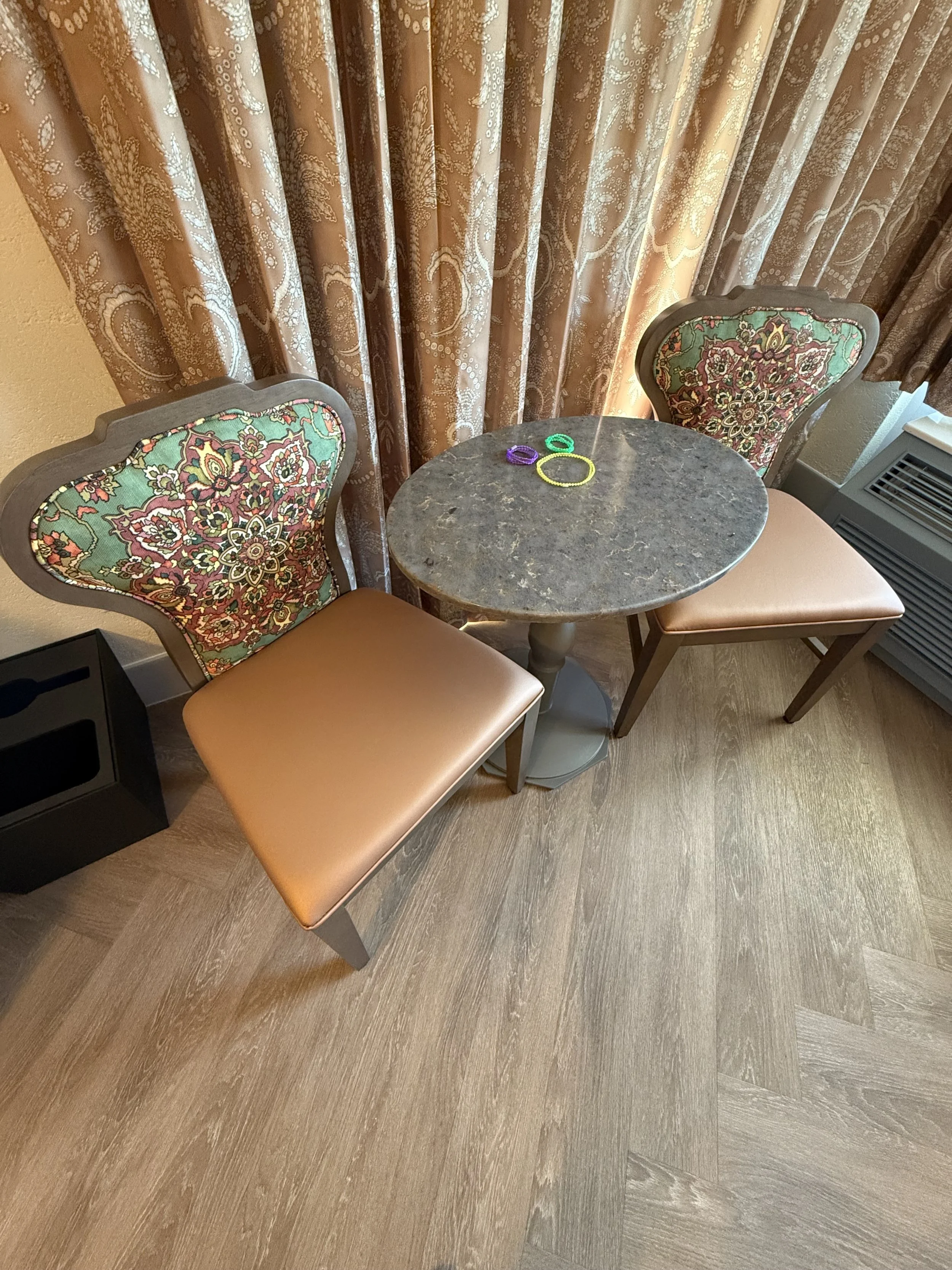 A small round marble table with two chairs, one with a floral upholstered back and the other with a plain padded seat. Several colorful rubber rings are on the table, set in front of a window with brown patterned curtains. The floor is light wood, an