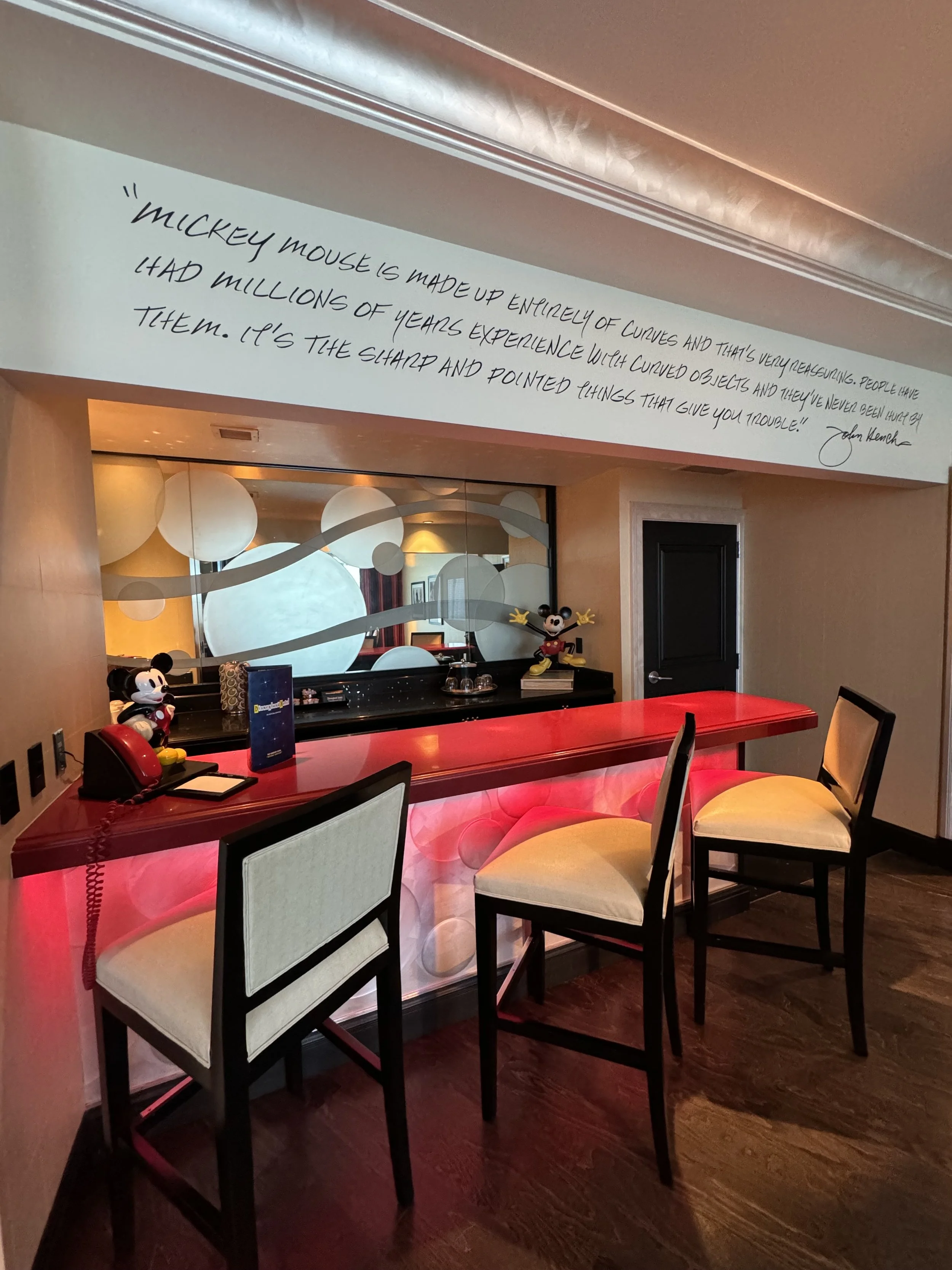 Decorative reception area with a red counter, Mickey Mouse figurines, a vintage telephone, and a quote about Mickey Mouse painted on the wall above.