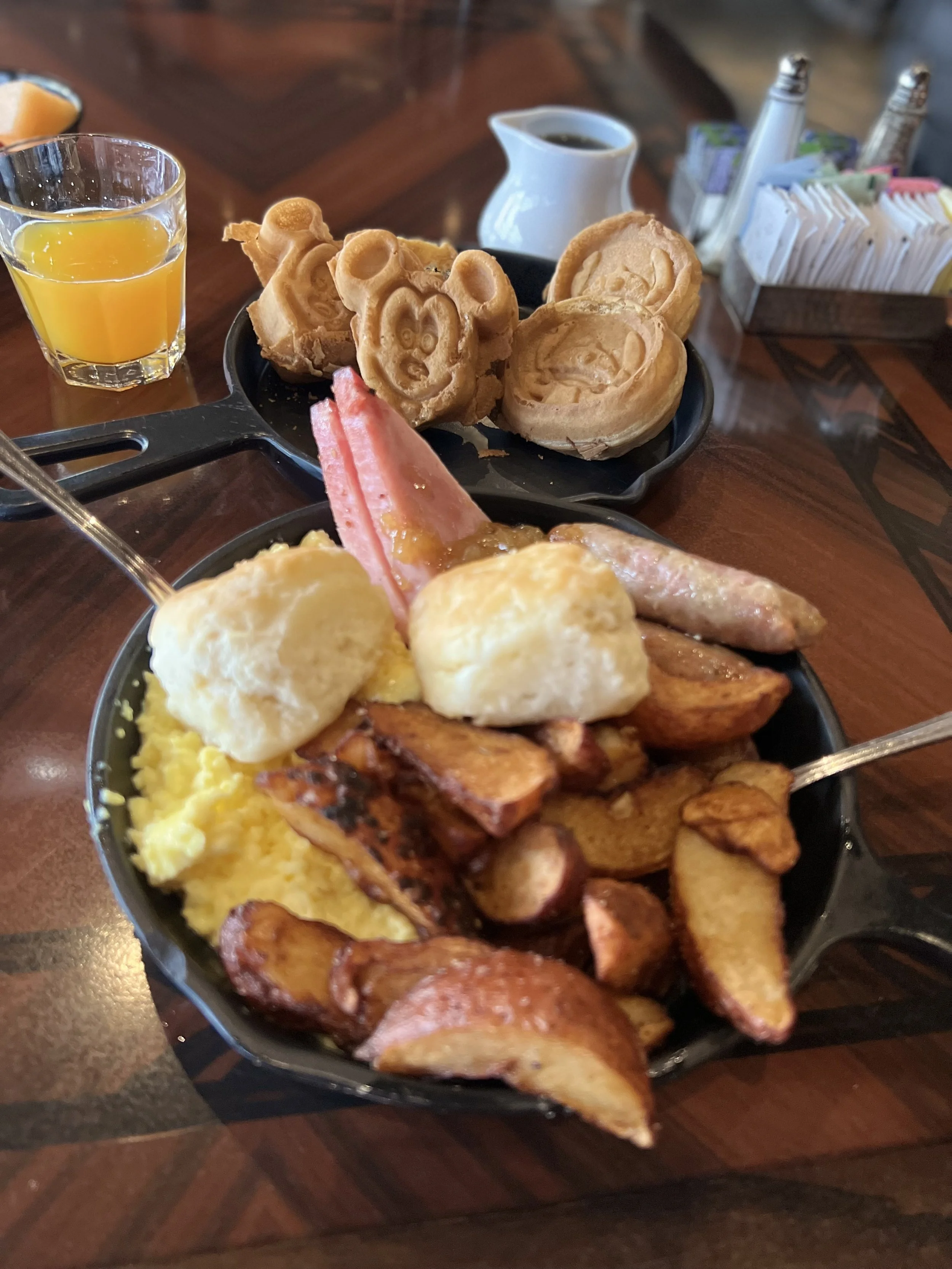A breakfast platter with scrambled eggs, bacon, sausage, and potato wedges. In the background, a plate of waffles with Mickey Mouse shapes and a cup of syrup.