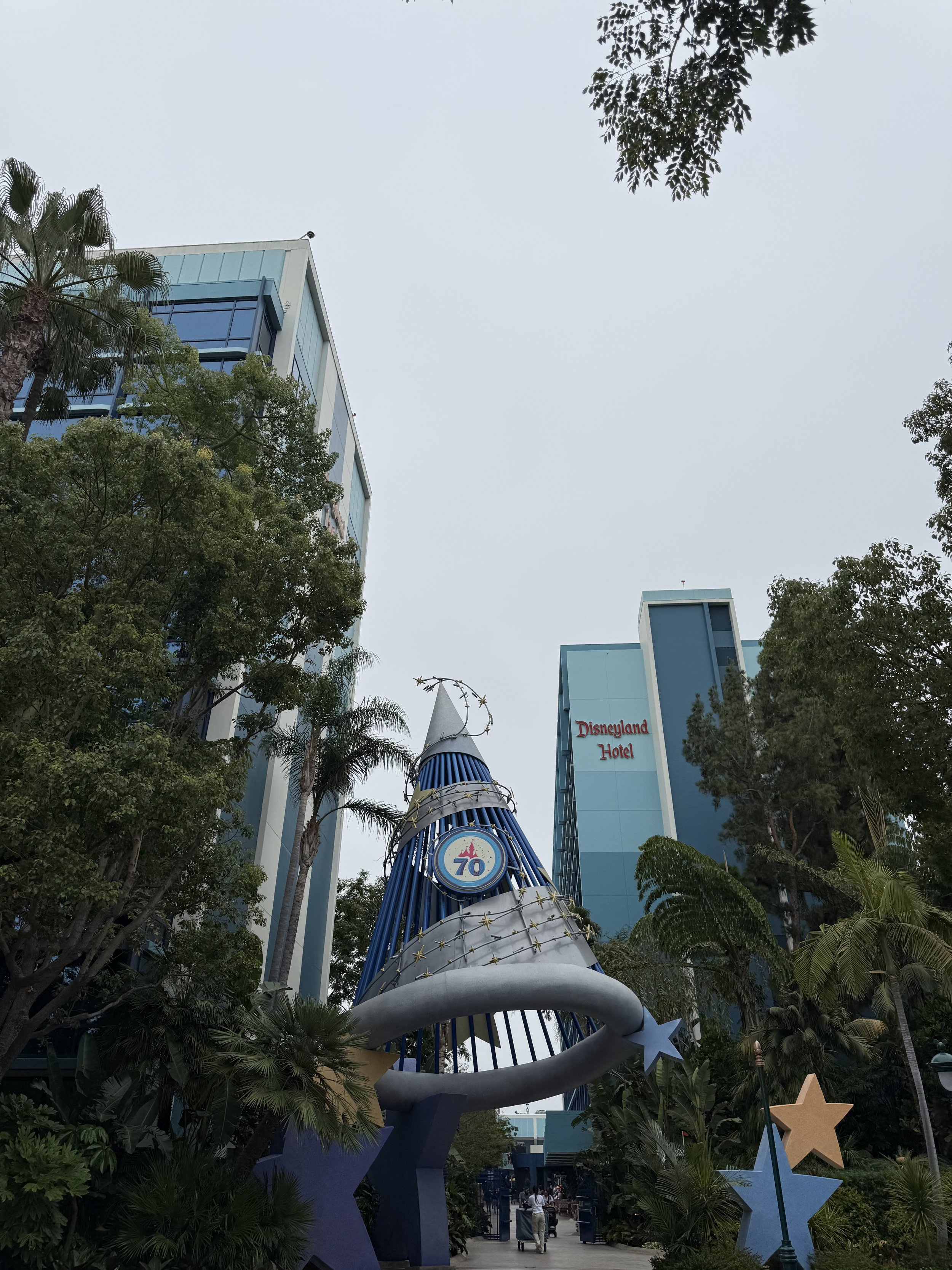 A Disney theme park entrance with a large birthday cake-themed decoration celebrating 70 years, surrounded by trees and tall blue buildings, including the Disneyland Hotel.