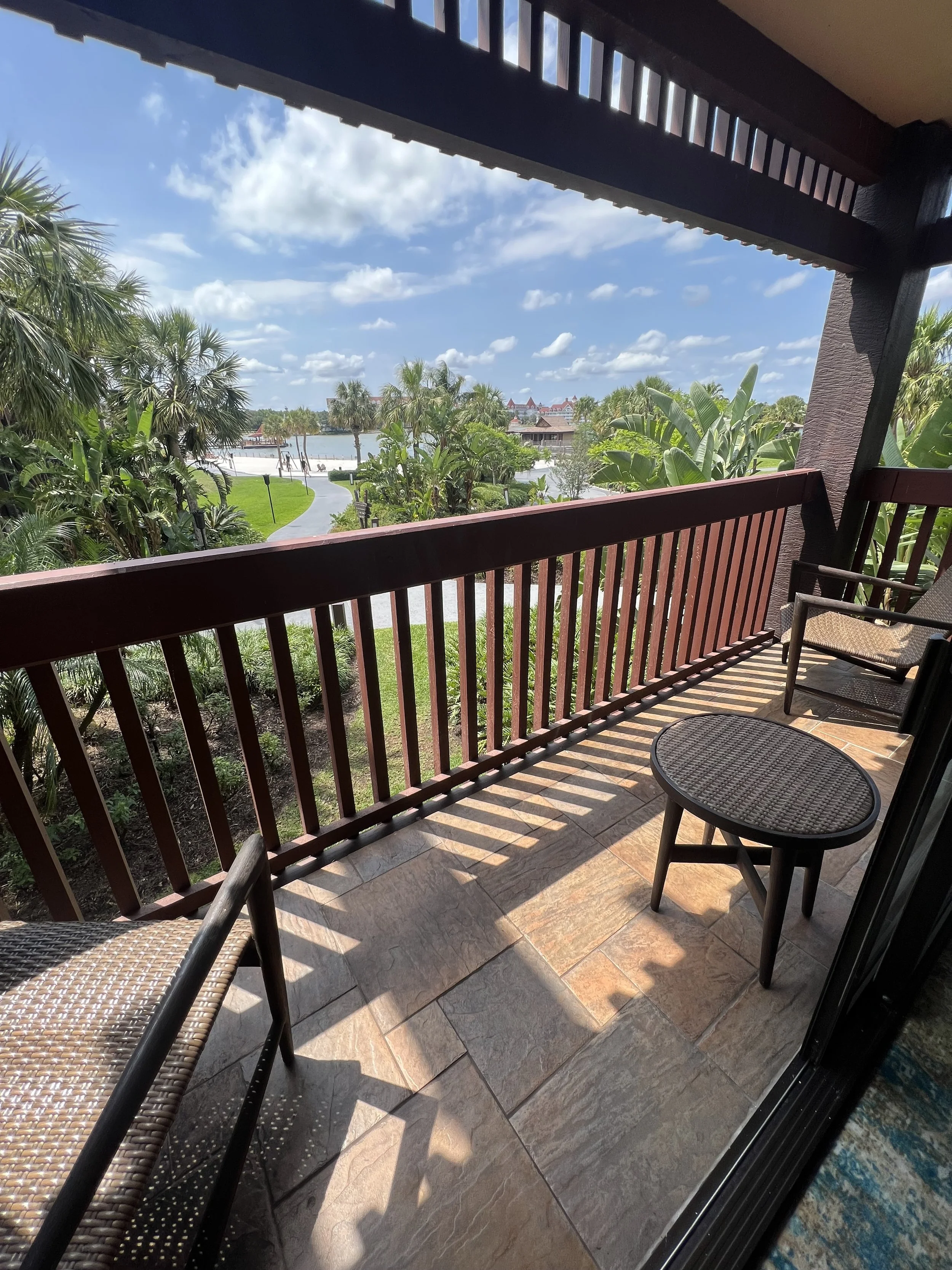 View from a balcony with wooden railing, two chairs, a small table, lush green palm trees and a pathway leading to a water body, under a cloudy blue sky.