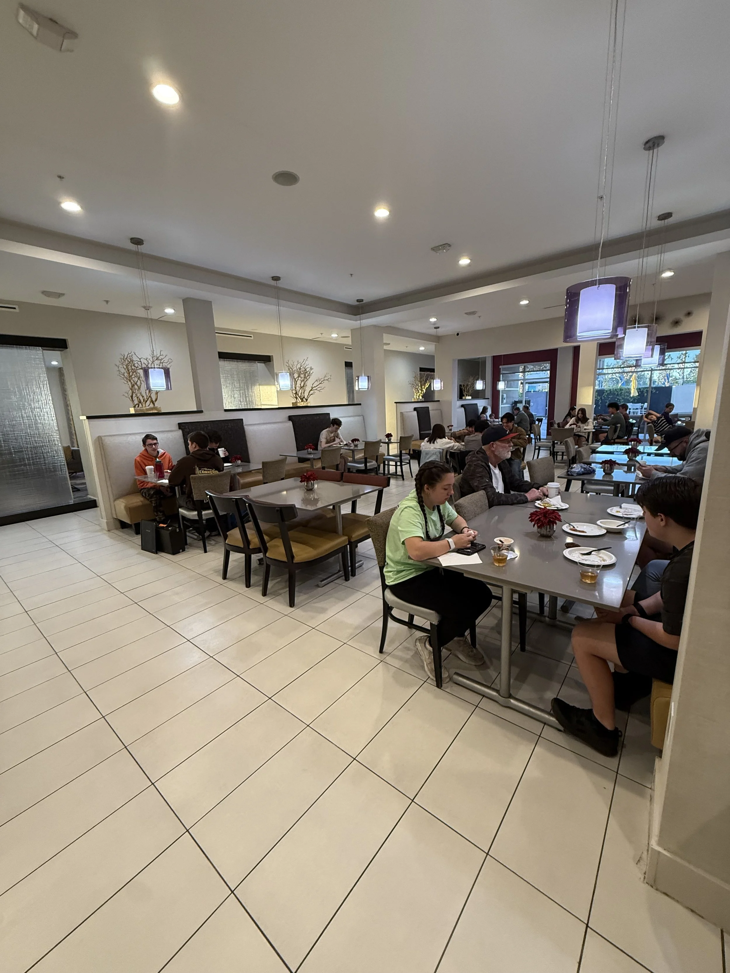 A busy restaurant with several groups of people dining at tables, decorated with flowers, large windows showing natural light, and modern lighting fixtures hanging from the ceiling.