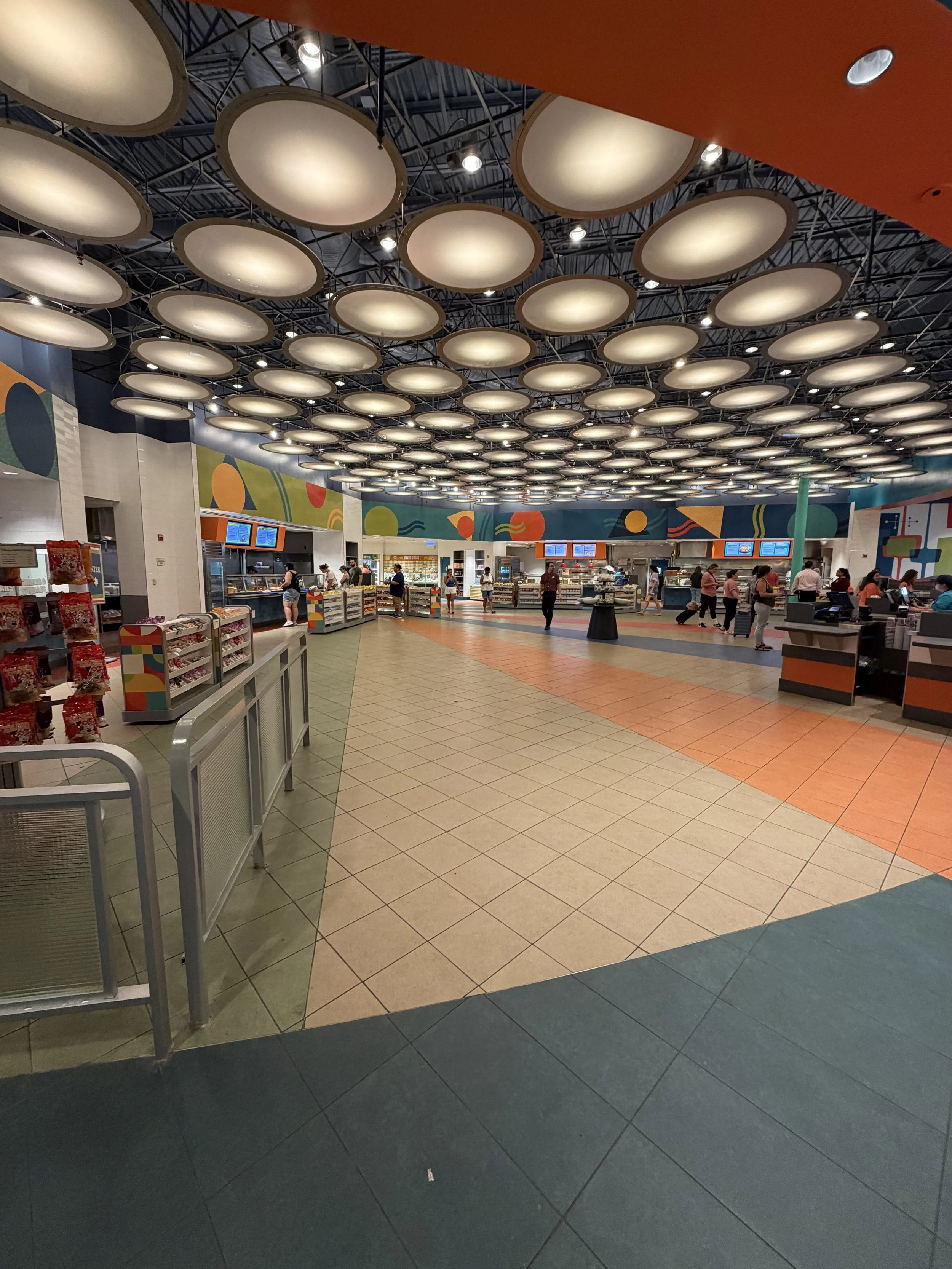 Inside a brightly lit, colorful cafeteria or cafeteria-style dining area with a patterned ceiling and a curved walkway. There are people standing in line and ordering food at multiple counters with digital menu boards, and shelves with snacks and oth