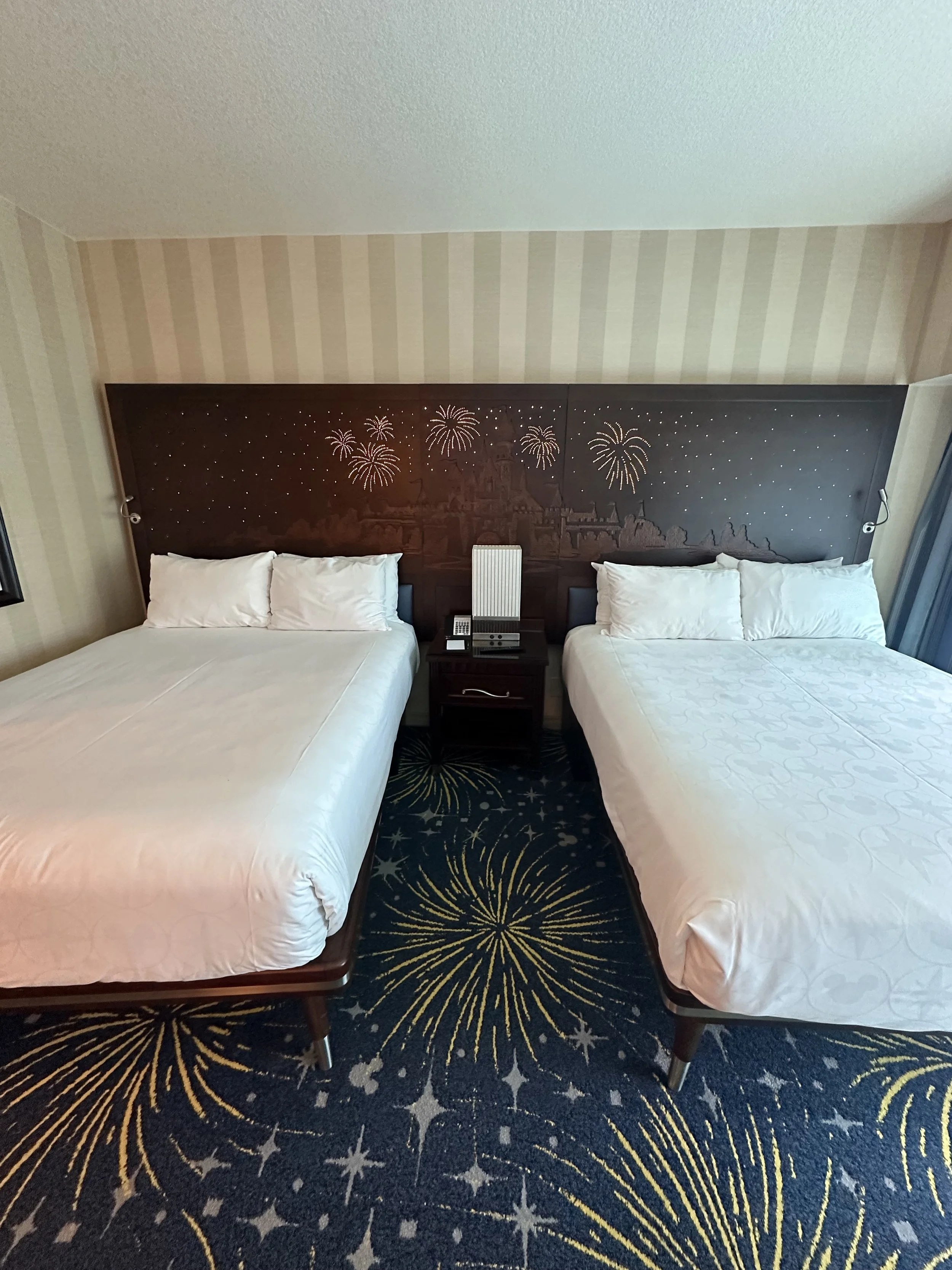 Two double beds with white linens and pillows in a hotel room, separated by a nightstand with a telephone and a speaker, with a decorated headboard featuring fireworks and castle designs, and firework patterns on the carpet.
