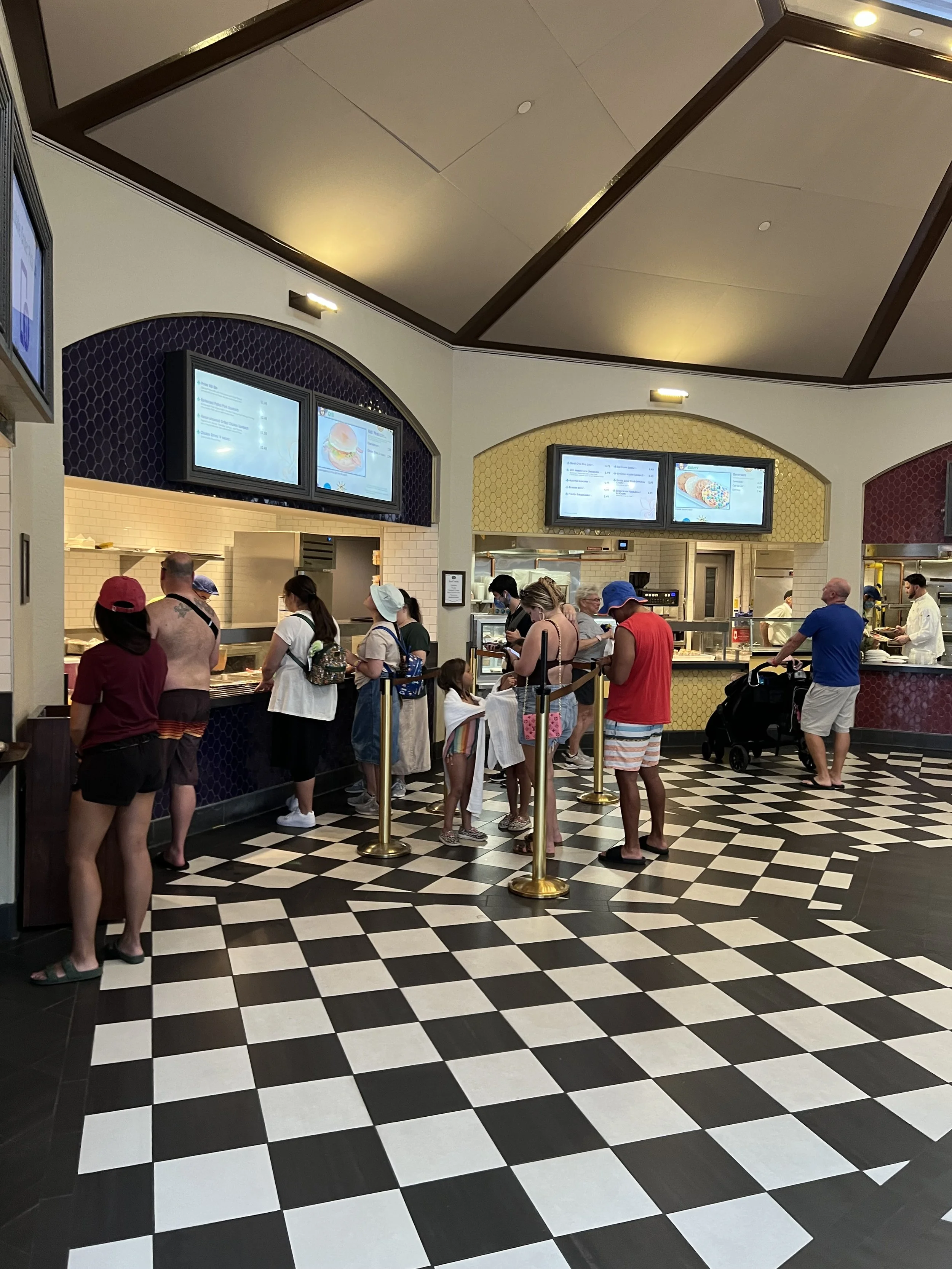 People waiting in line at a counter in a restaurant with digital menu screens overhead, checkered black and white floor, and a kitchen in the background.