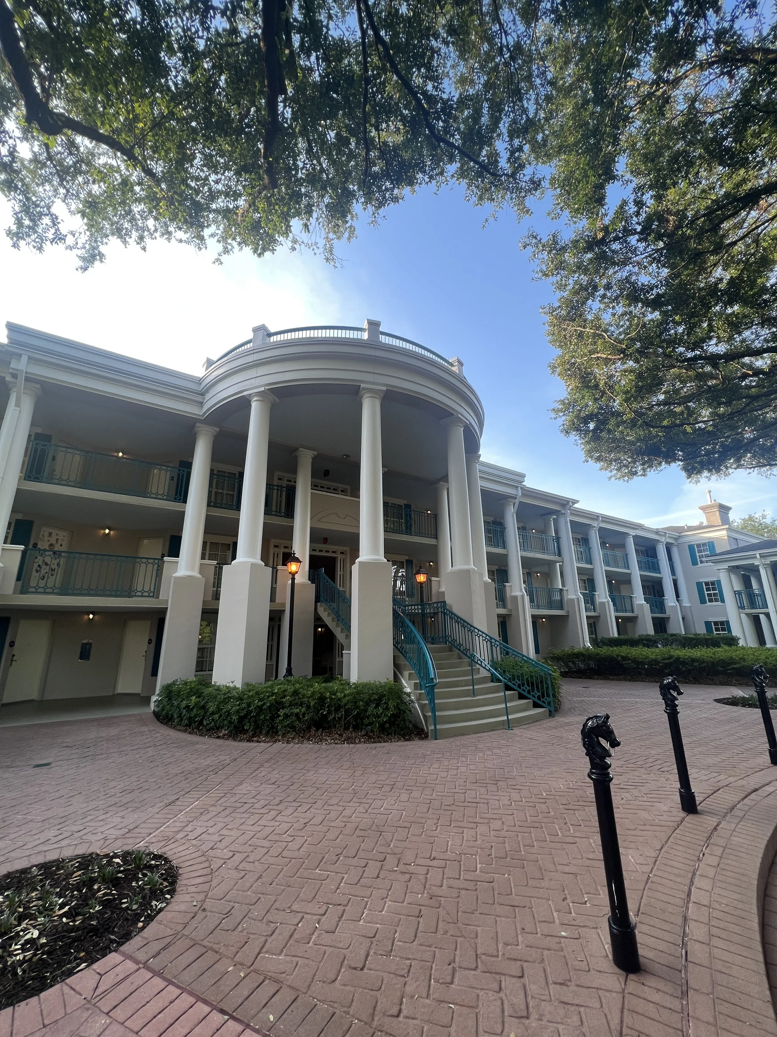 A three-story white building with large columns and a curved staircase leading to the entrance. It features balconies with blue railings, surrounded by trees and a brick-paved walkway. There are lamp posts near the stairs and decorative horse head bo