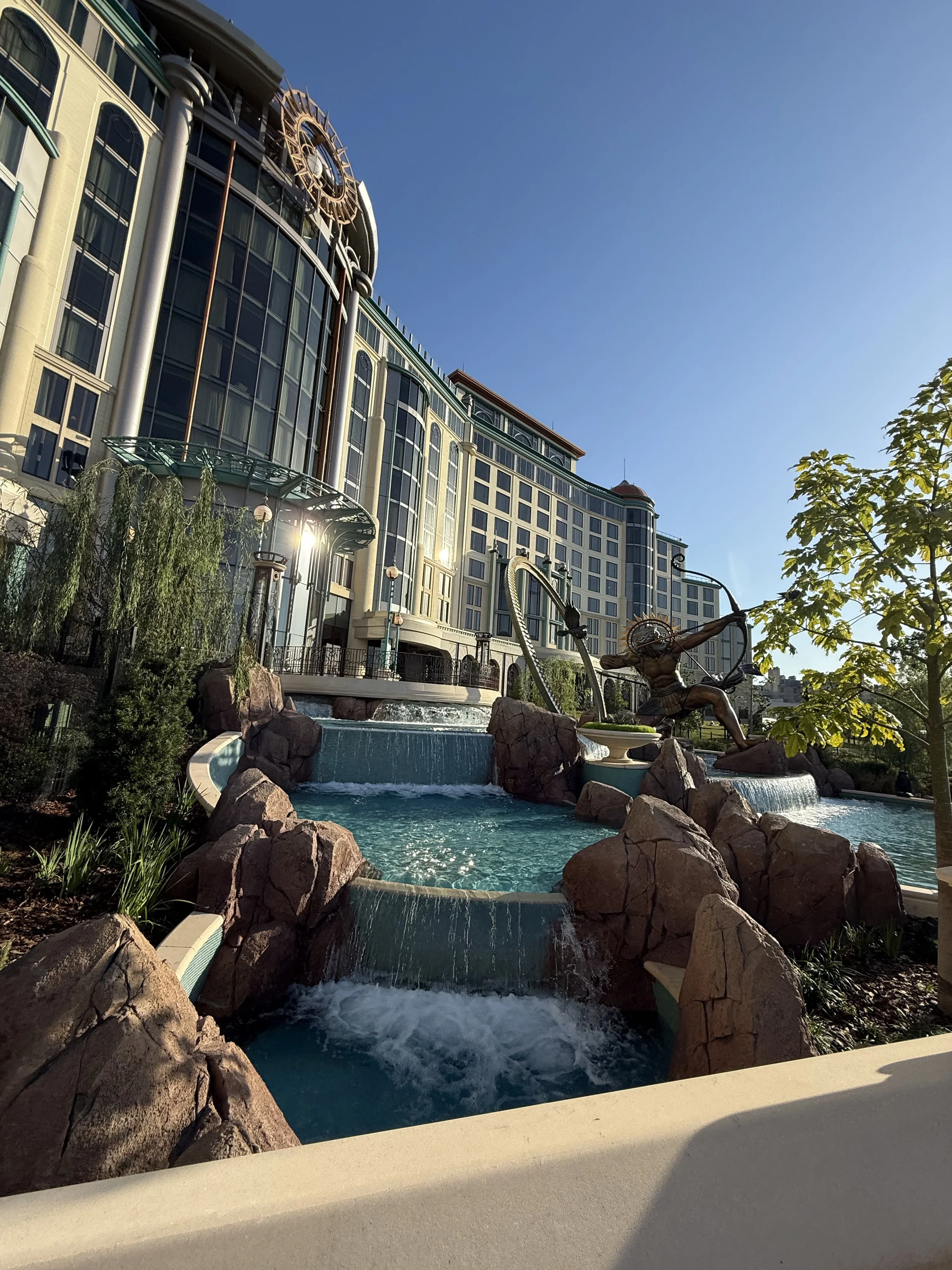 A large hotel or casino building with a water fountain and statue of a woman with a bow in front, surrounded by rocks and greenery, under a clear blue sky.