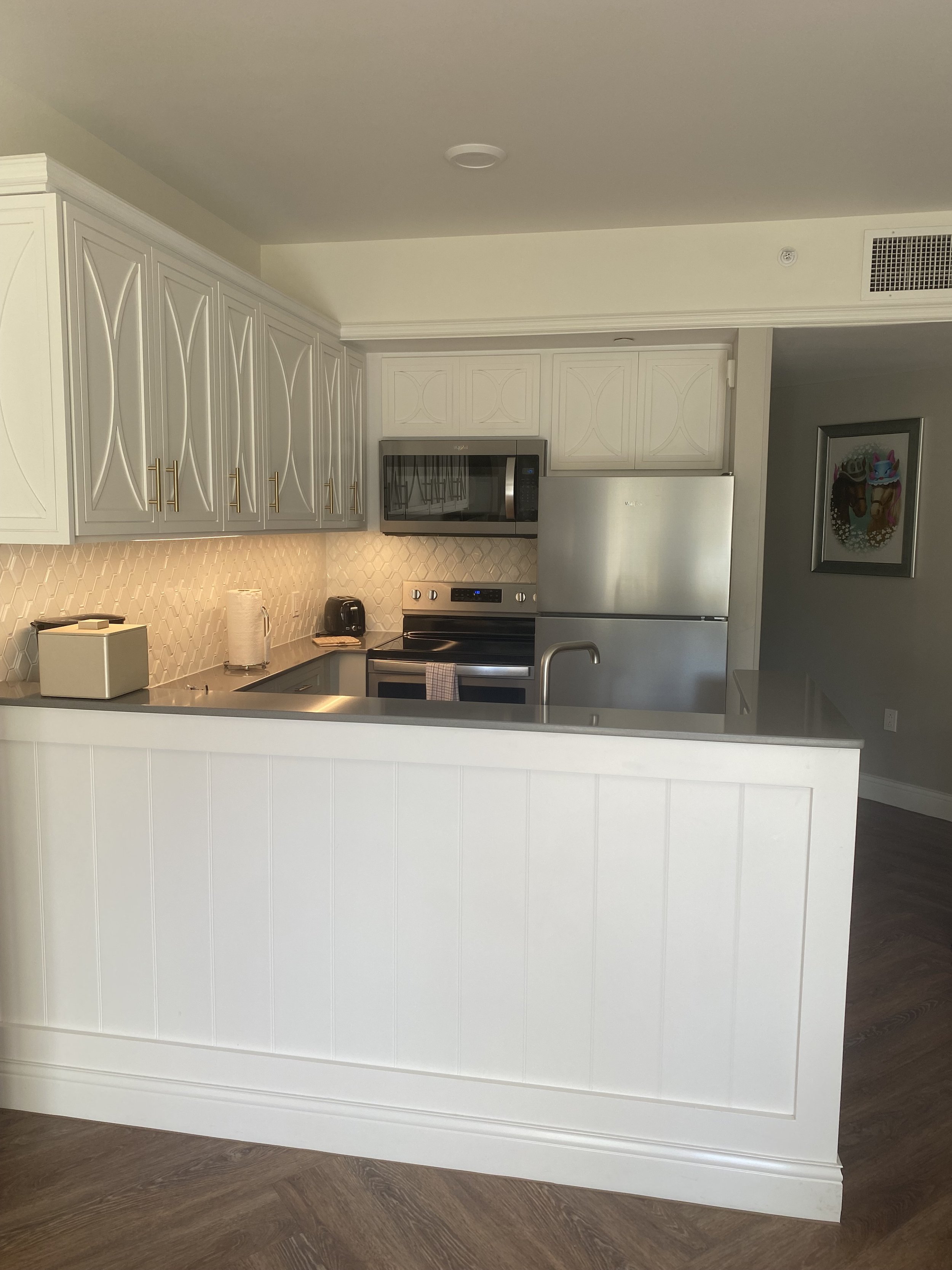 Kitchen with white cabinets, gray countertops, stainless steel refrigerator, microwave, and oven, with a white backsplash and some small appliances on the counter.