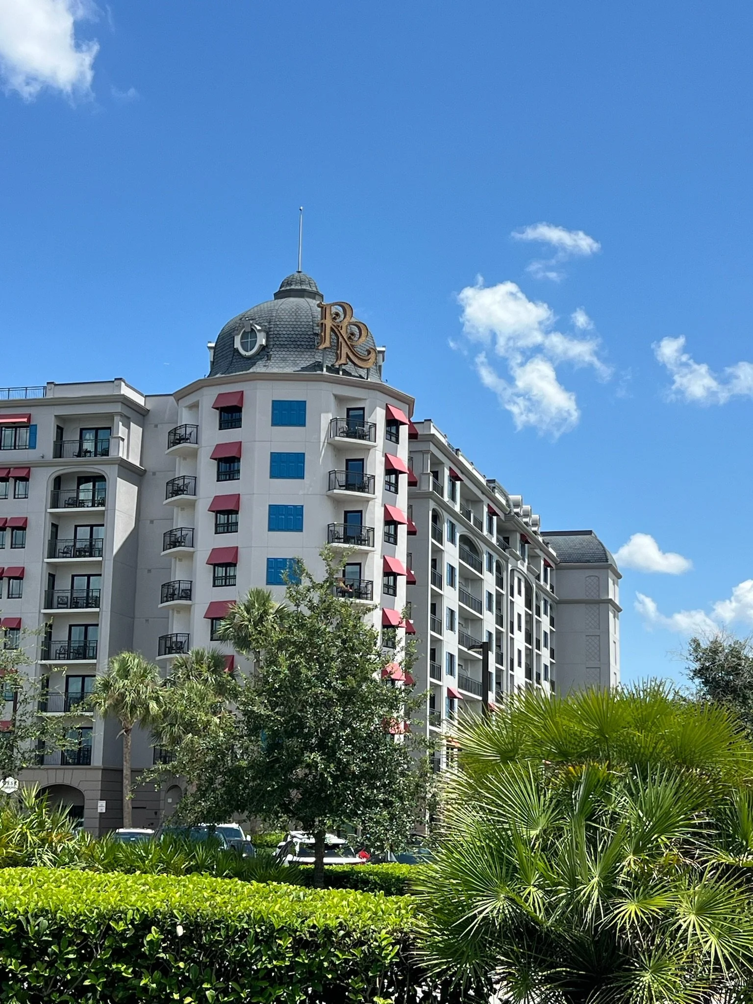 A multi-story building with a domed top, red awnings, and balconies, surrounded by greenery and palm trees under a blue sky with scattered clouds.