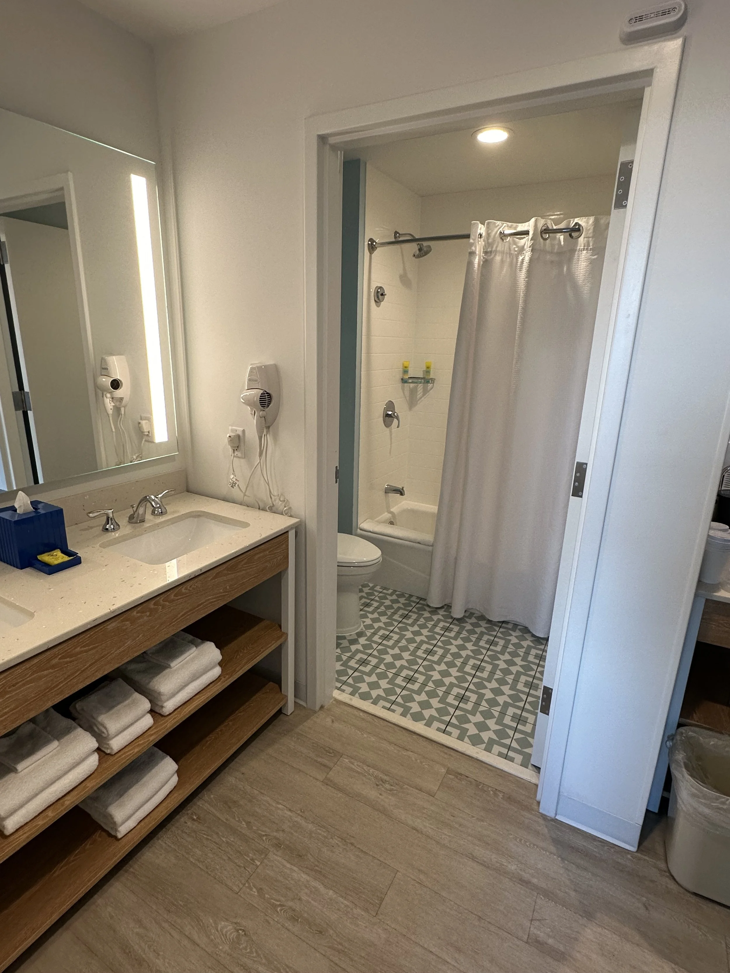 A bathroom with a vanity sink on the left, towels on open shelves underneath, a mirror with lights, and a wall-mounted hairdryer. In the background, a doorway leads to a shower and bathtub area with a white curtain, a showerhead, soap holder, and two