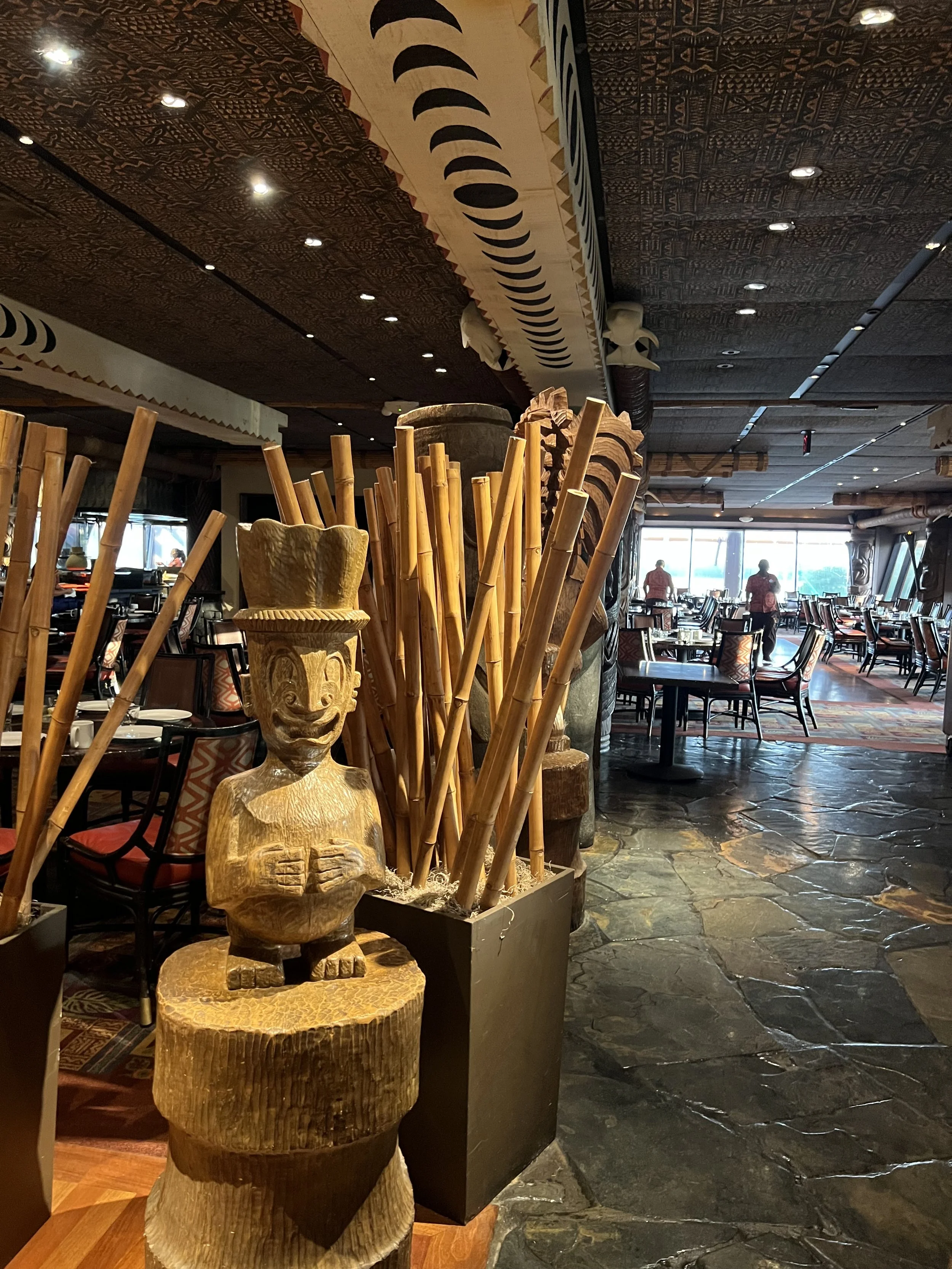 Decorative wooden carving of a smiling figure with a hat and hands on chest, surrounded by bamboo sticks, inside a restaurant with tables and chairs, and large windows with a view outside.