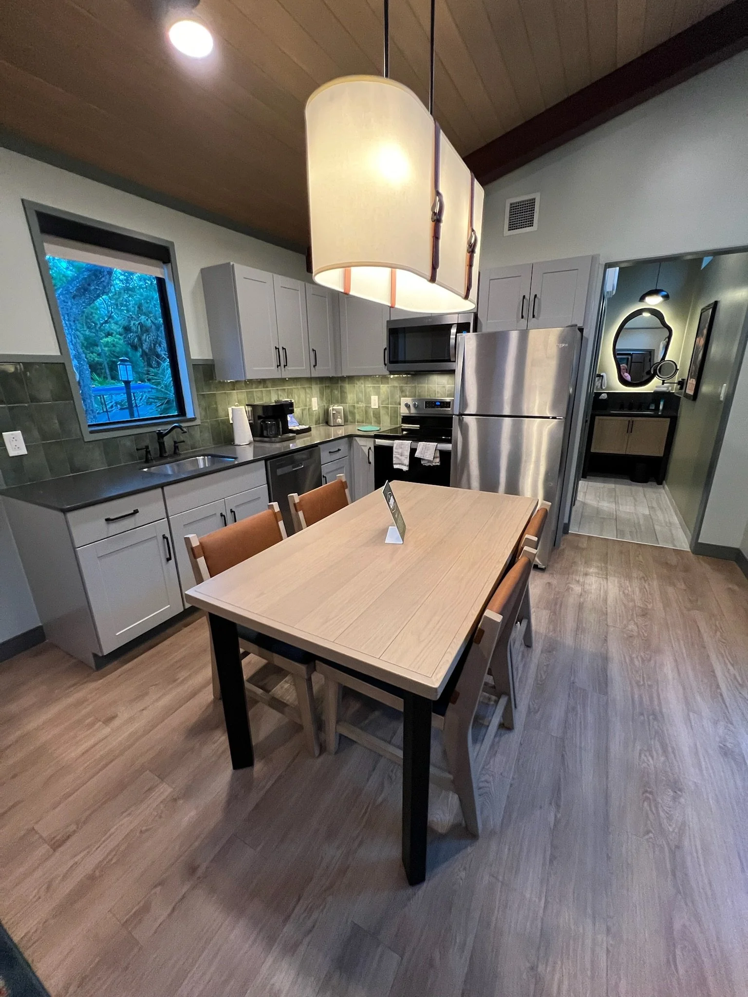 Kitchen with a wooden dining table and six chairs, stainless steel refrigerator, microwave, oven, green tiled backsplash, window showing outdoor greenery, and a hanging ceiling light.