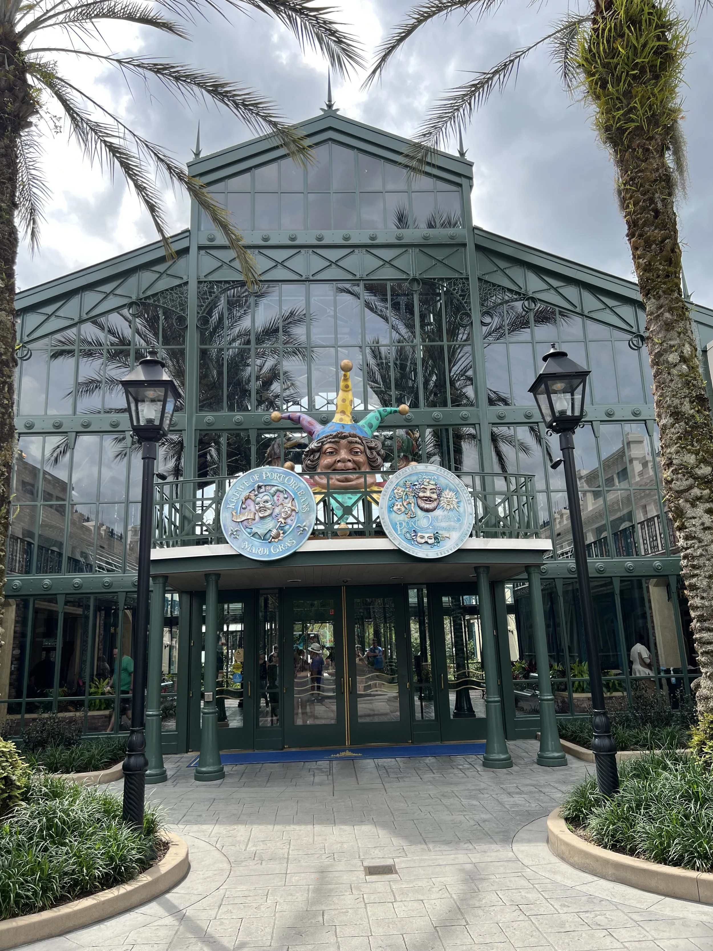 Greenhouse with a large jester face sculpture wearing a colorful hat. Two circular signs hang on the balcony, and there are palm trees on either side of the building. People are visible inside and outside, and the area is landscaped with plants and a