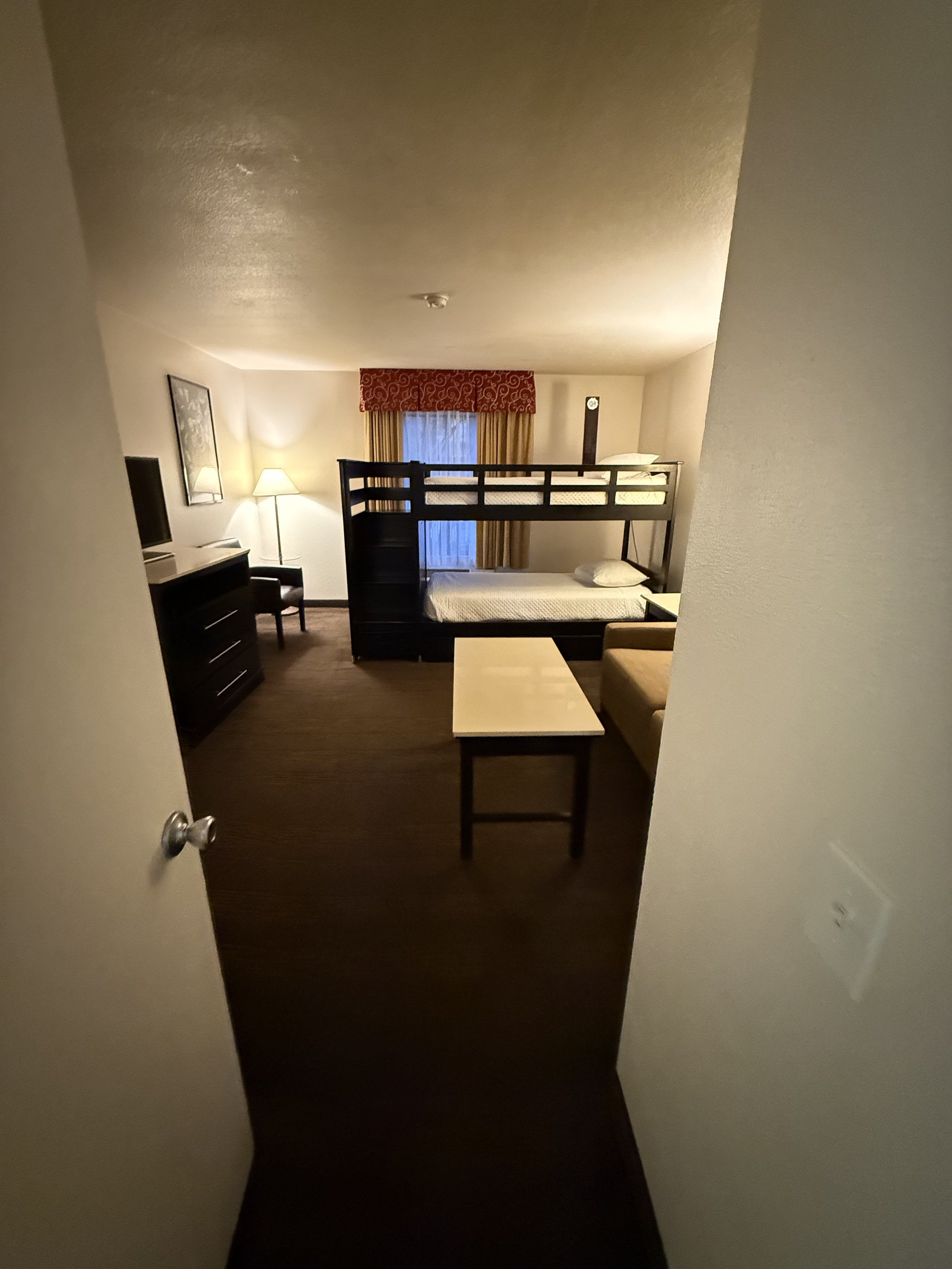 Hotel room with bunk beds, a dresser with a television, a chair, a small table, a sofa, and a window with curtains.