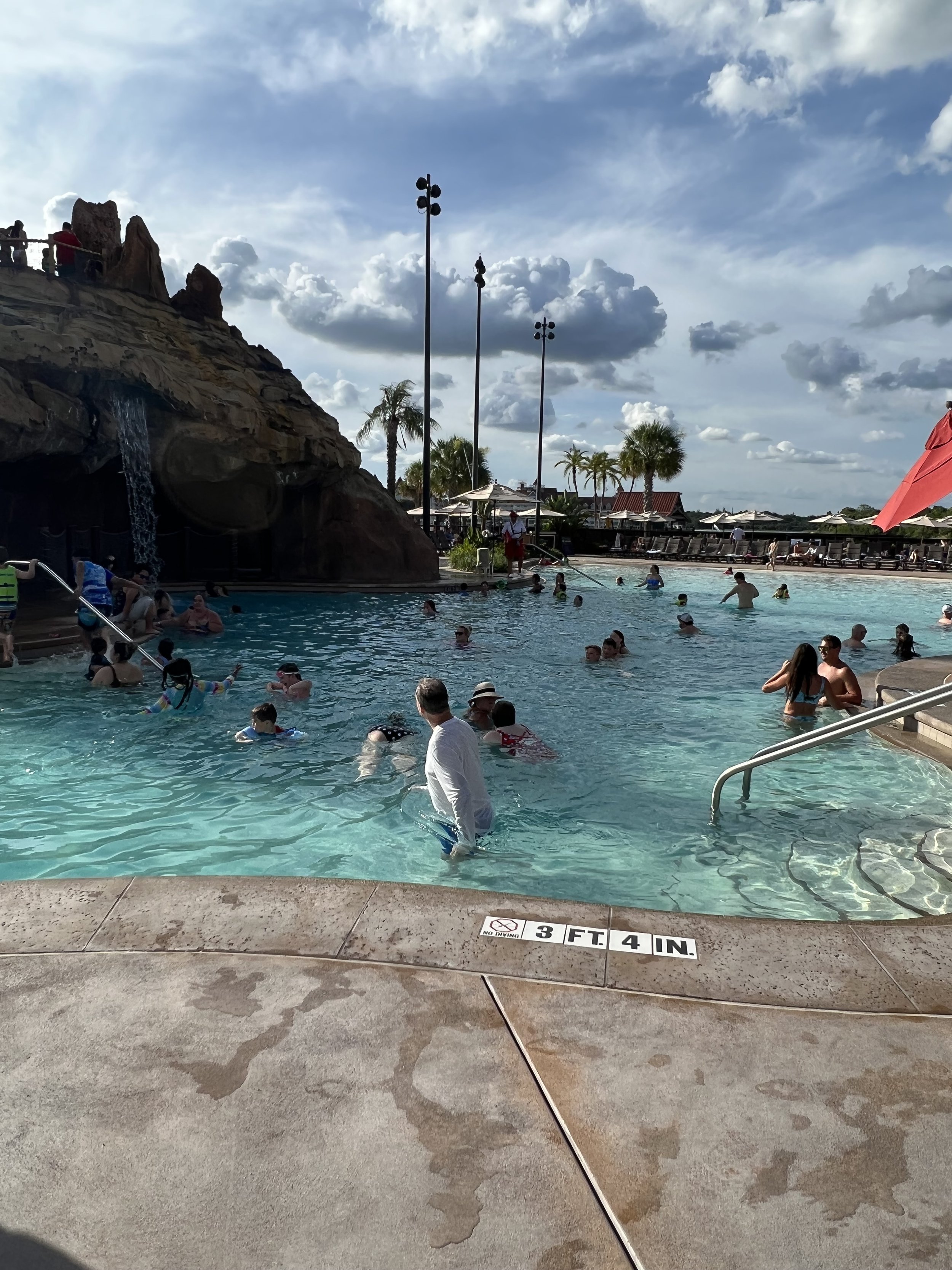 People swimming and relaxing at a pool with artificial rock waterfall, palm trees, lounge chairs, and umbrellas under a partly cloudy sky.