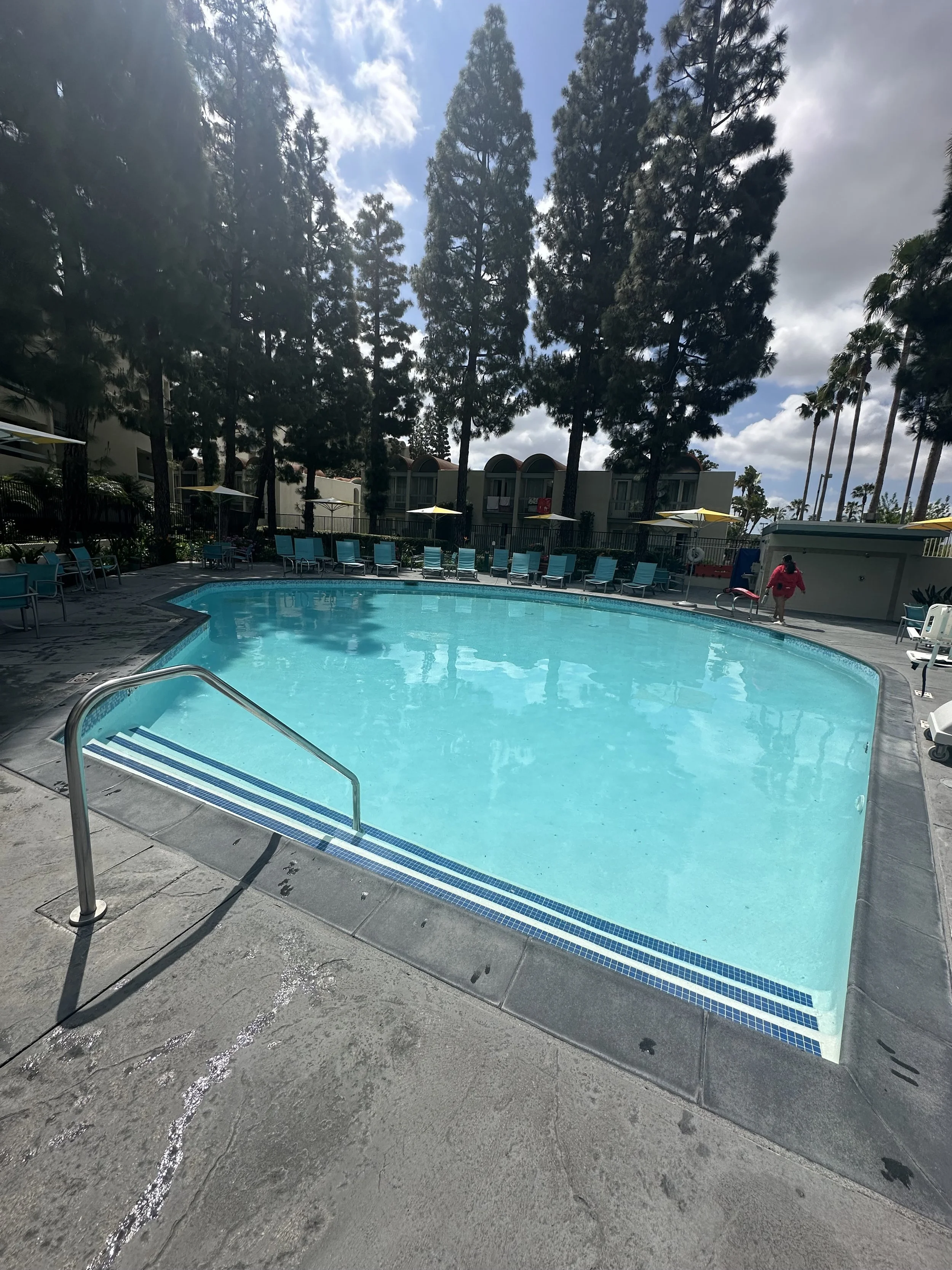 Empty outdoor swimming pool with blue water and a metal handrail, surrounded by blue lounge chairs and yellow umbrellas, tall trees, apartment buildings in the background, partly cloudy sky.