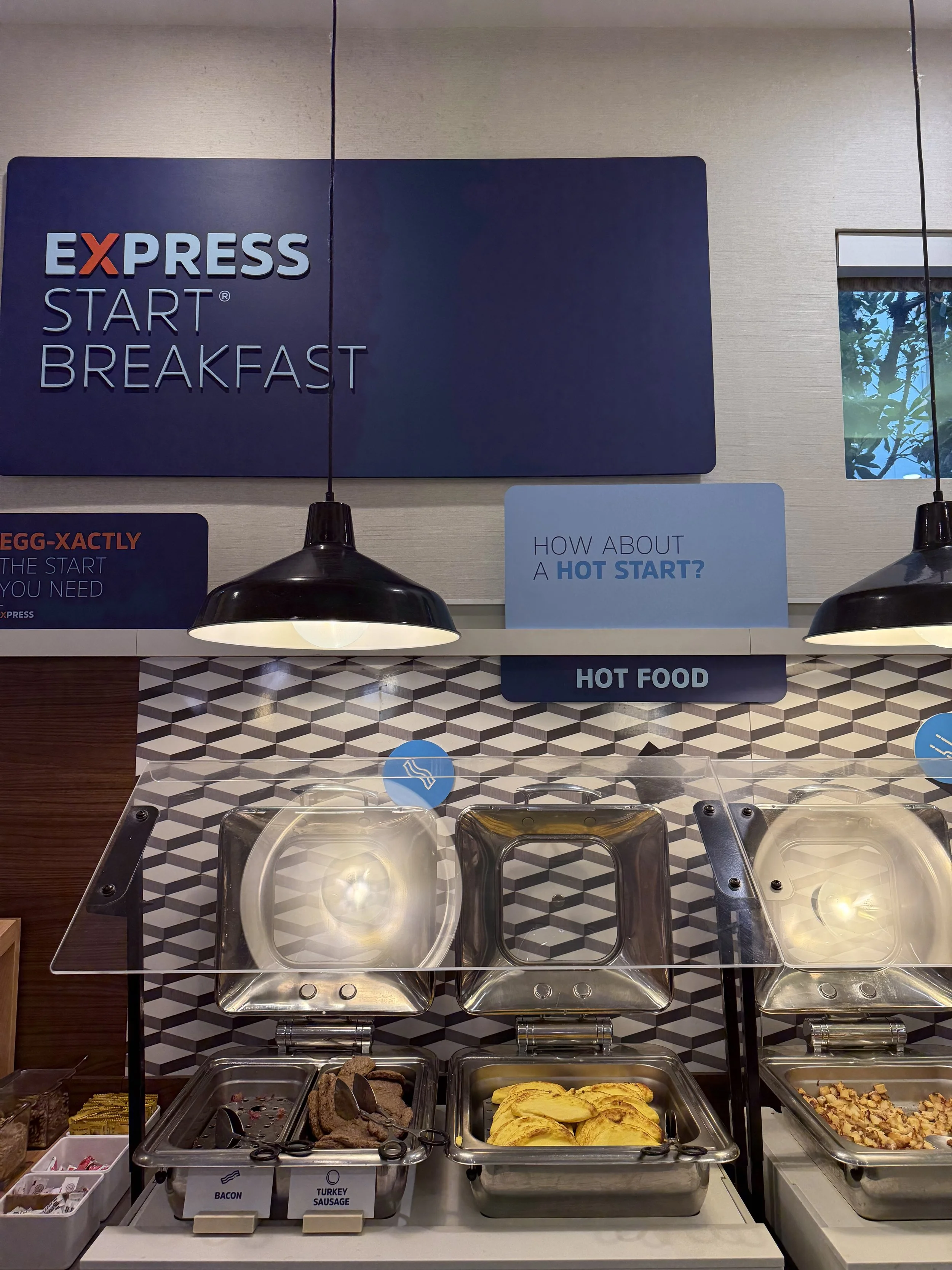 Breakfast buffet at a fast food restaurant with trays of bacon, turkey sausage, and scrambled eggs, under a sign that reads 'Express Start Breakfast' and a smaller sign asking about a hot start.