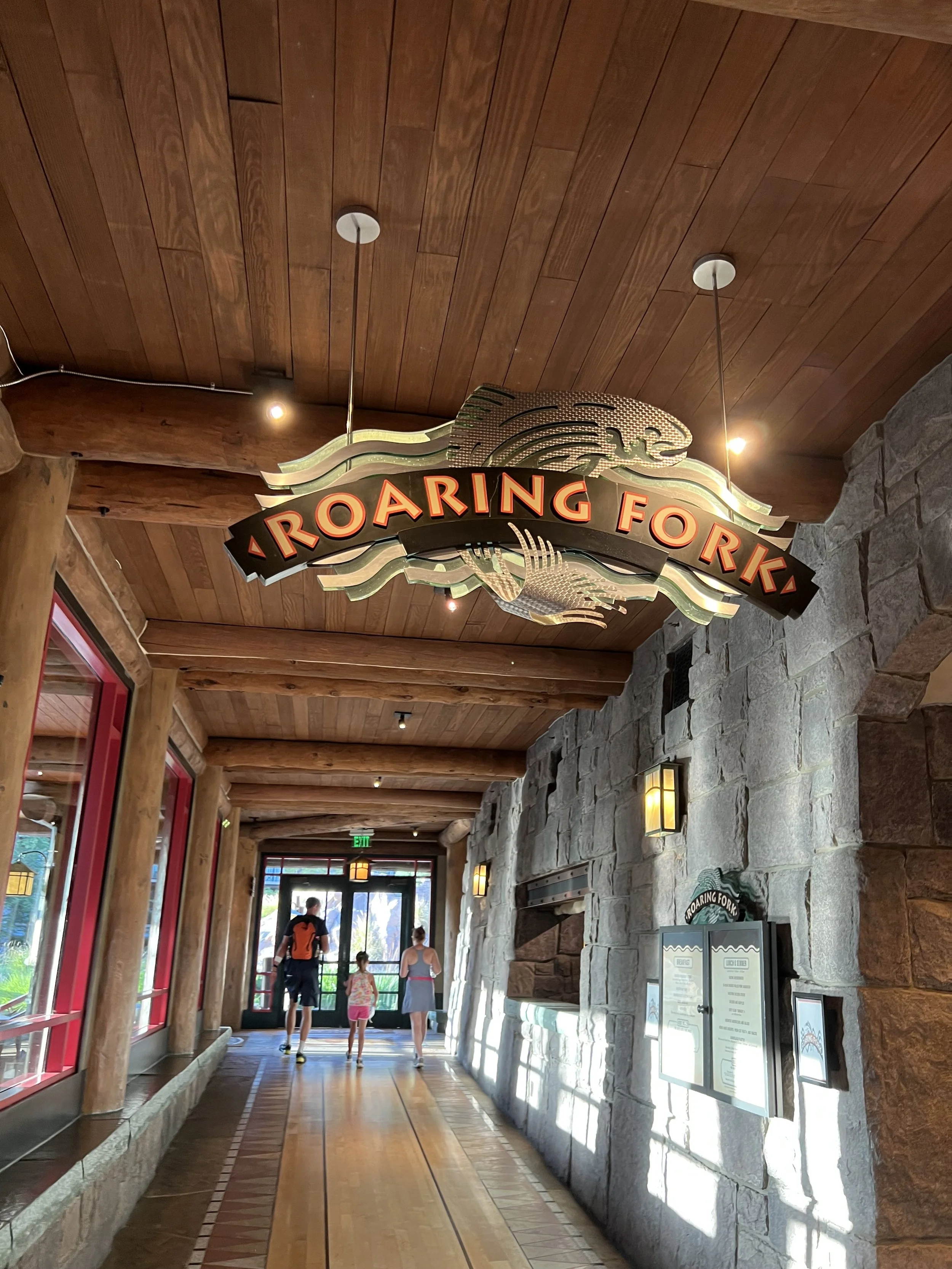 Interior hallway of a zoo or wildlife park with a sign that reads 'Roaring Fork,' showing a mountain lion or cougar graphic. The hallway has wooden ceilings, stone walls, and large windows, with a family walking towards a door at the end.