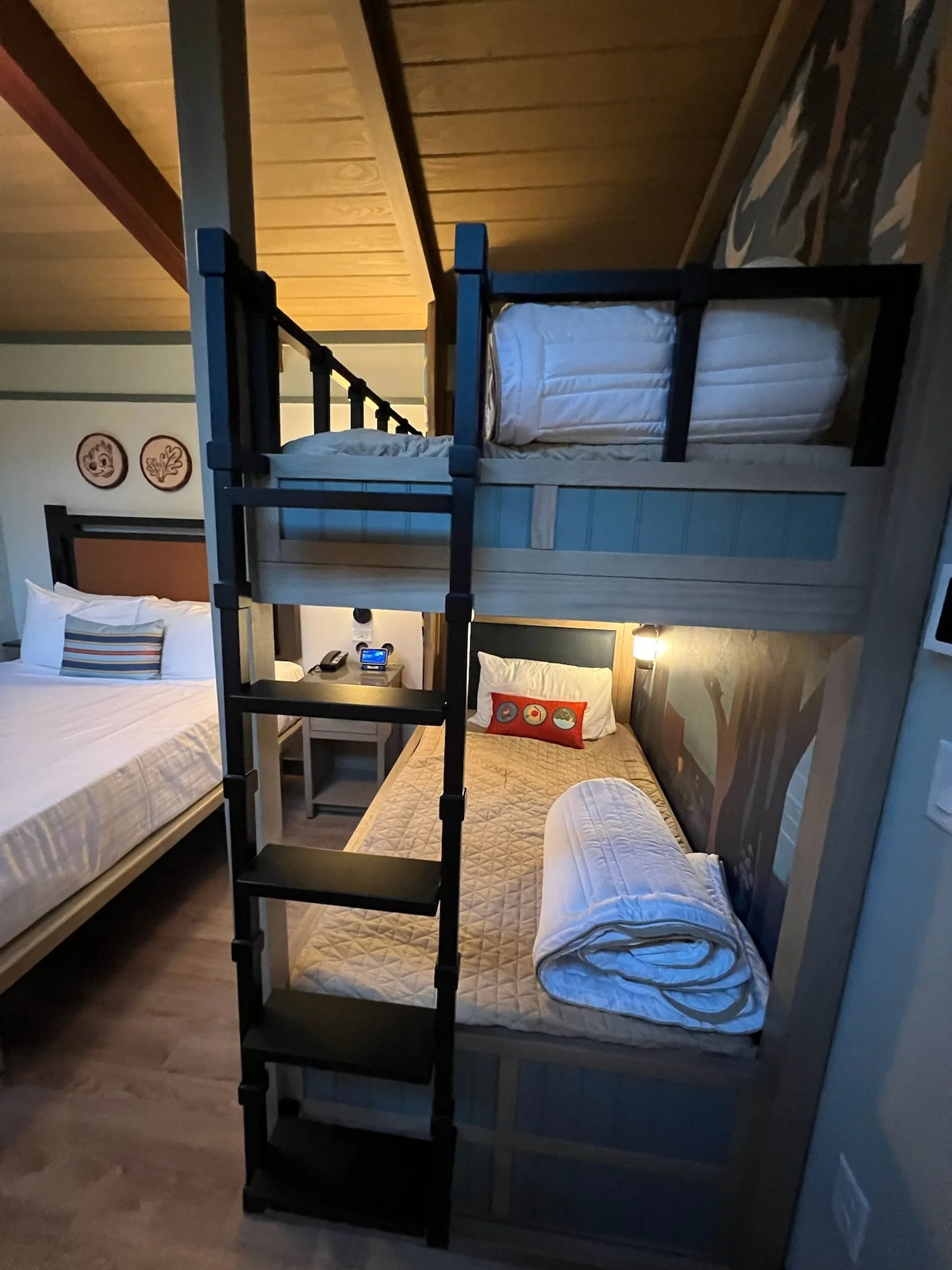 A bedroom with a bunk bed above a single bed, with rolled-up towels on the lower bed and a pillow with a colorful design. There are two beds, and the room has wooden walls and ceiling, with a nightstand and a lamp between the beds.