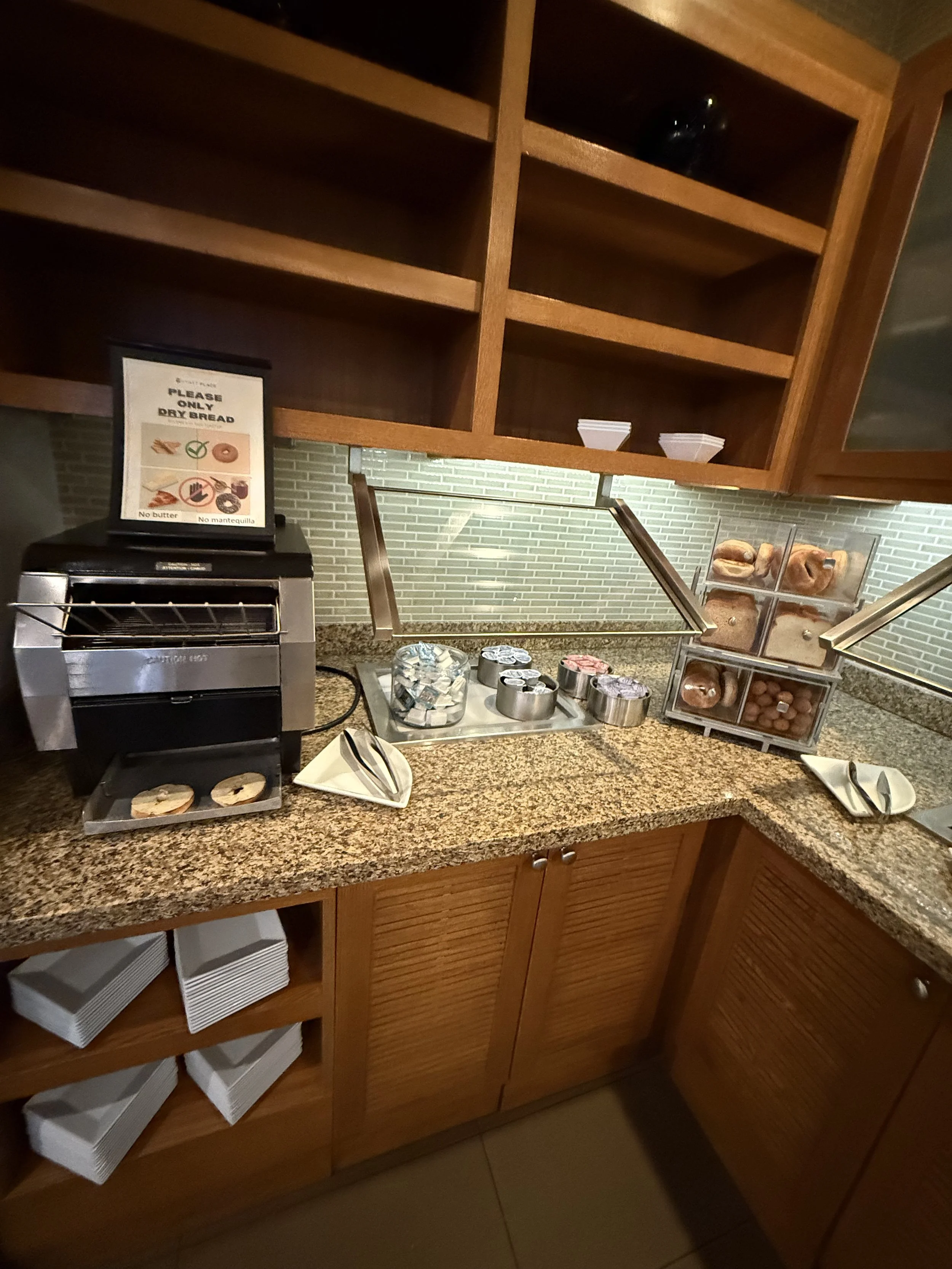 Deli counter with bread and snacks, including bagels and muffins, in a hotel or restaurant setting with a sign requesting only dry bread, a toaster, and empty shelves above.