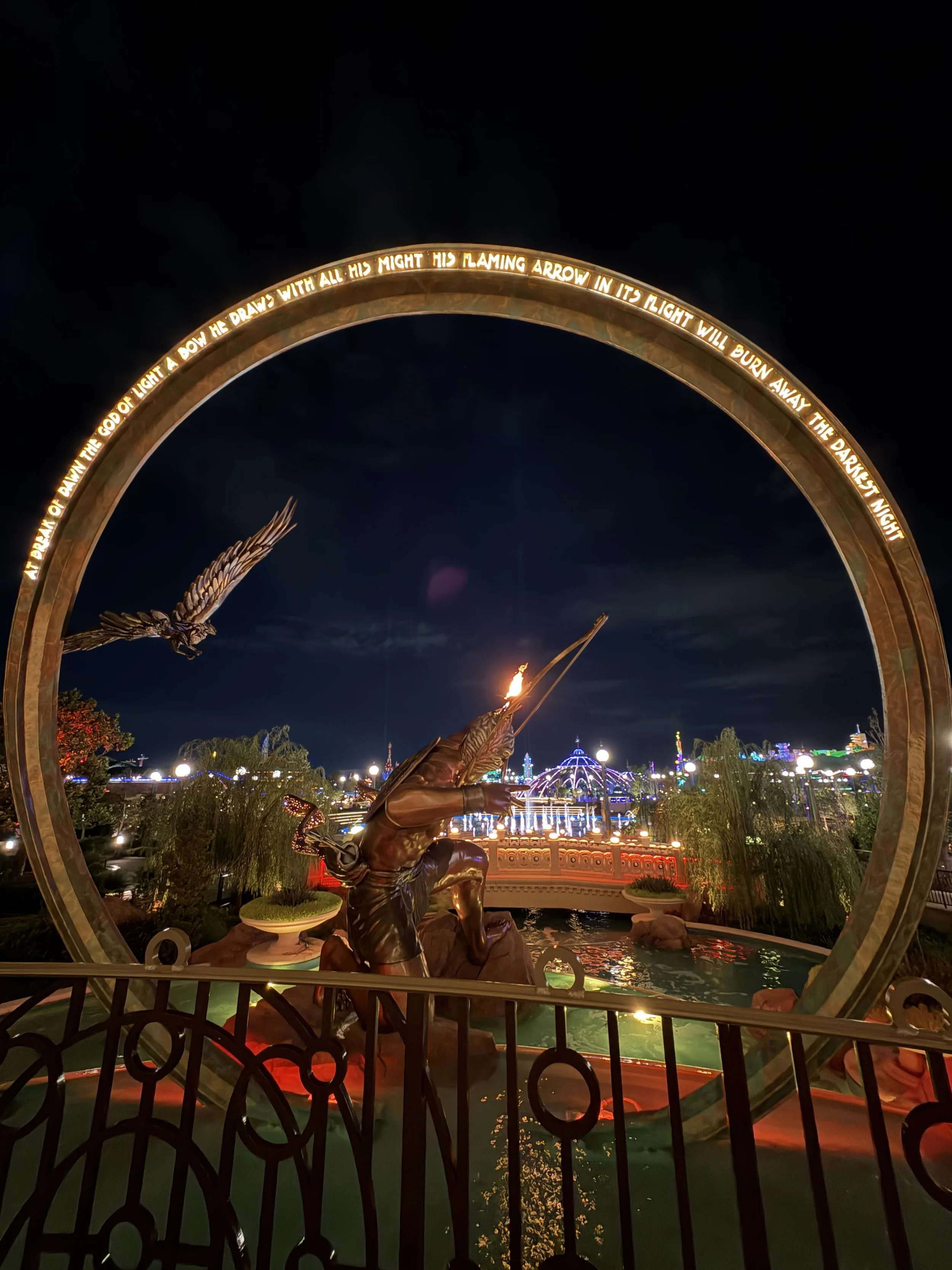 Statue of a Native American warrior aiming a flaming arrow at night, with a circular arch with engraved text surrounding it, and illuminated amusement park rides in the background.