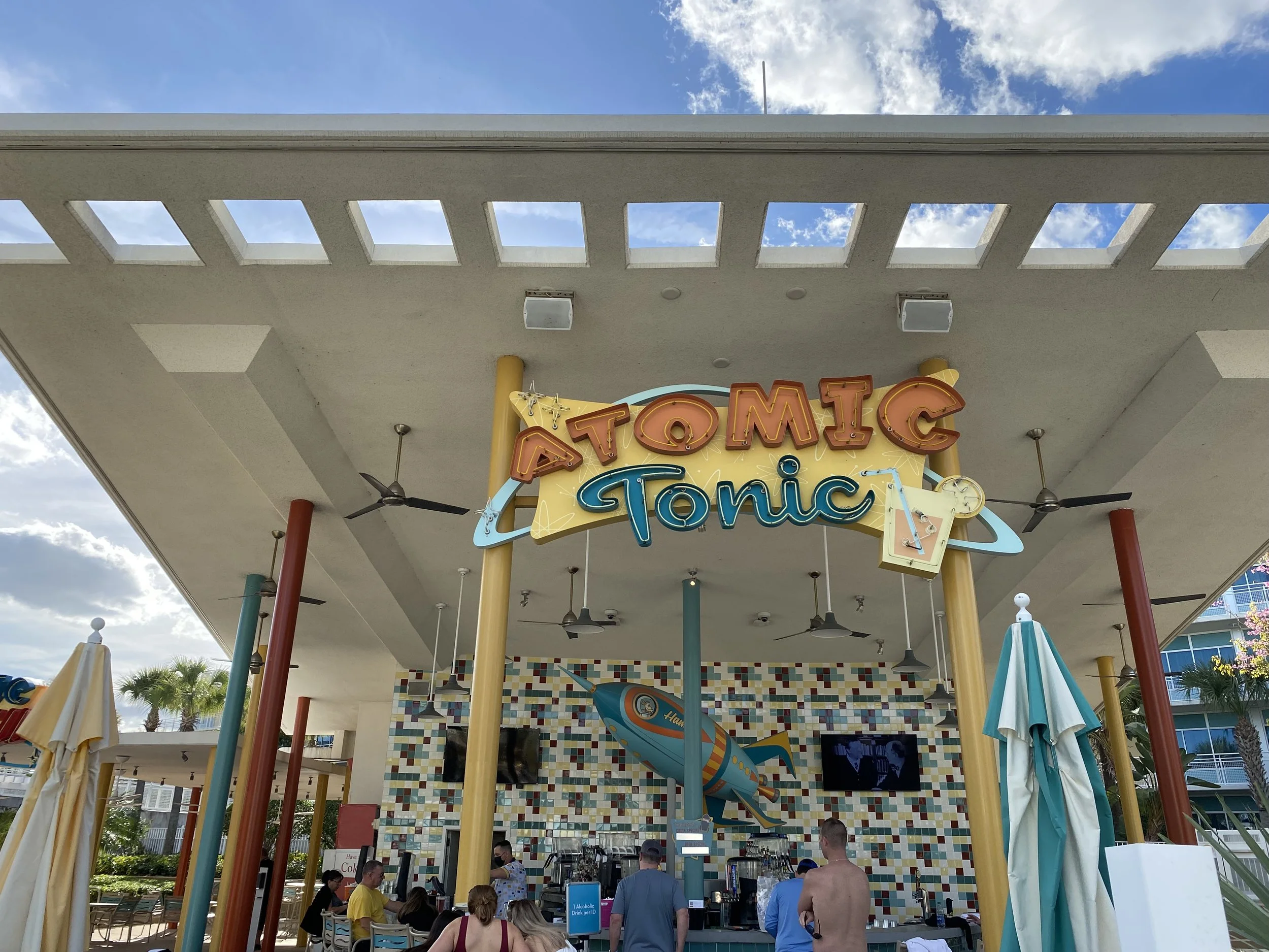 Outdoor bar area with a sign that reads 'Atomic Tonic,' featuring a colorful, vintage-style rocket and a yellow, blue, and red color scheme. Several people are gathered at the bar, and there are ceiling fans and umbrellas present.