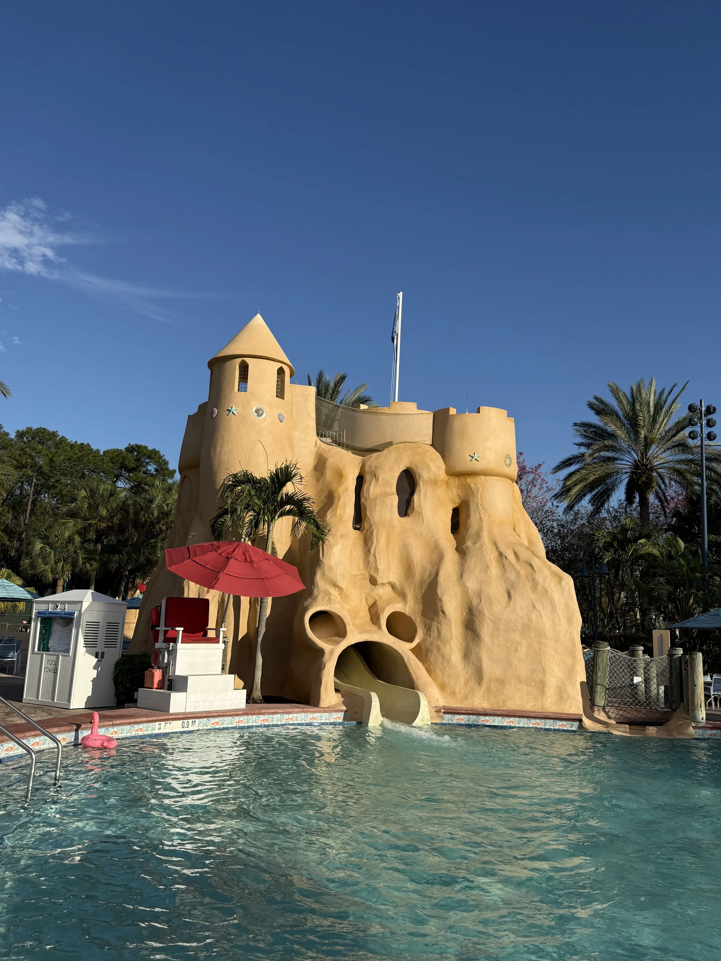 A colorful swimming pool with a yellow castle-shaped water slide, a pink flamingo float, and green palm trees under a clear blue sky.