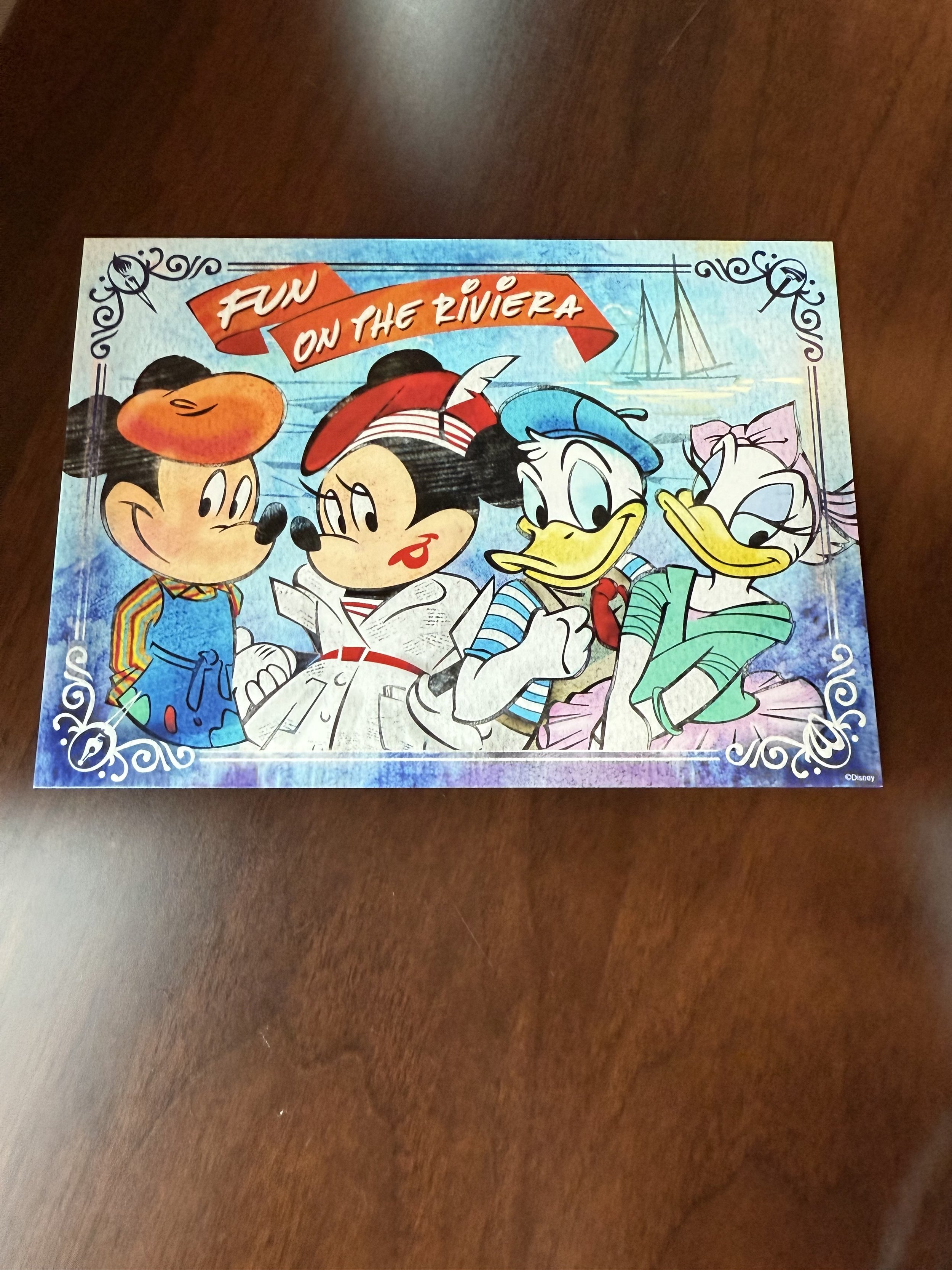 Colorful illustrated postcard featuring Disney characters Mickey Mouse, Minnie Mouse, Donald Duck, and Daisy Duck, with a background of water and sailboats, and a banner reading 'Fun on the Riviera.'