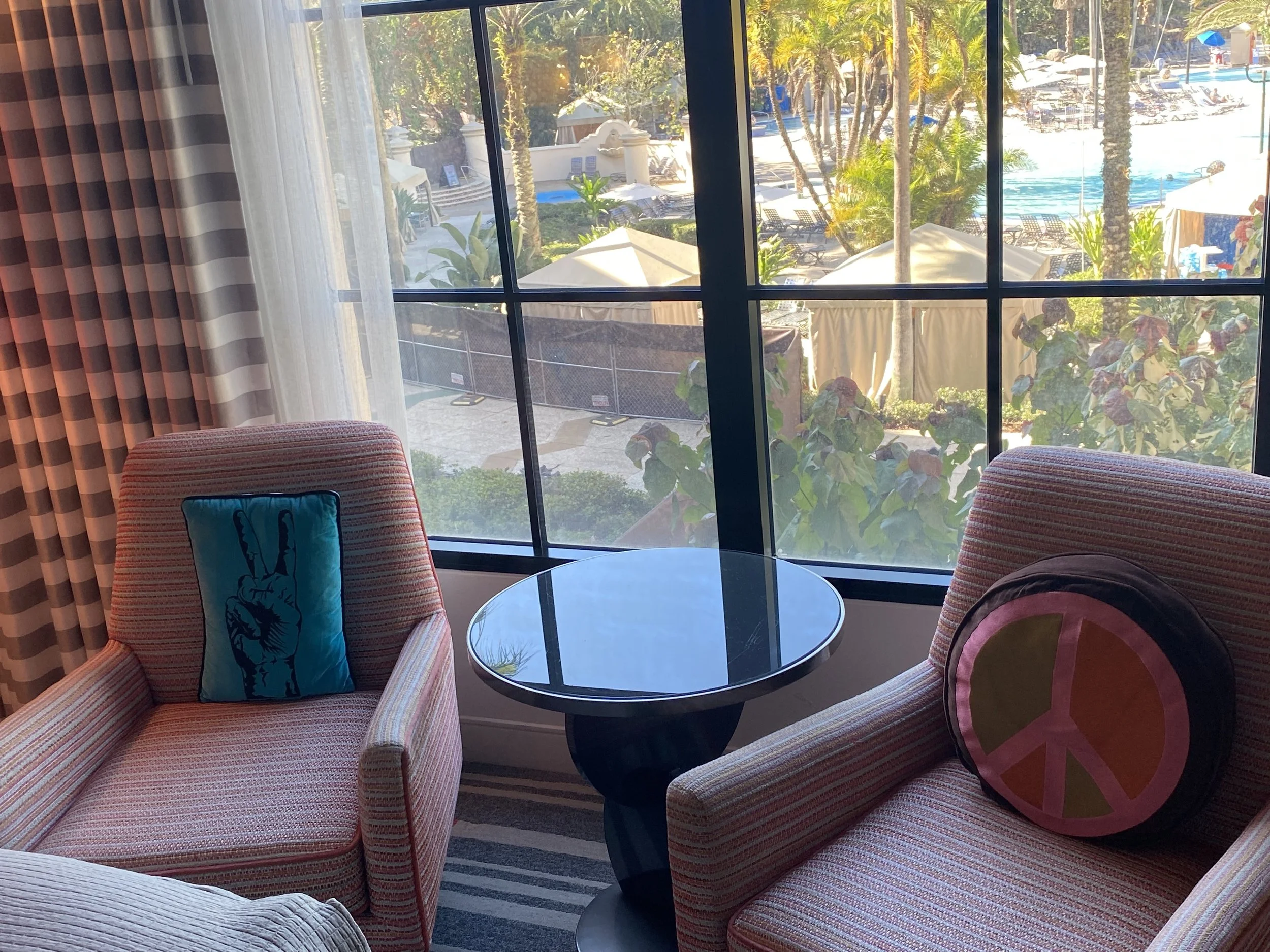 Seating area with two striped armchairs, pink and yellow pillows with peace symbol, round glass table in front, large window overlooking pool and palm trees.