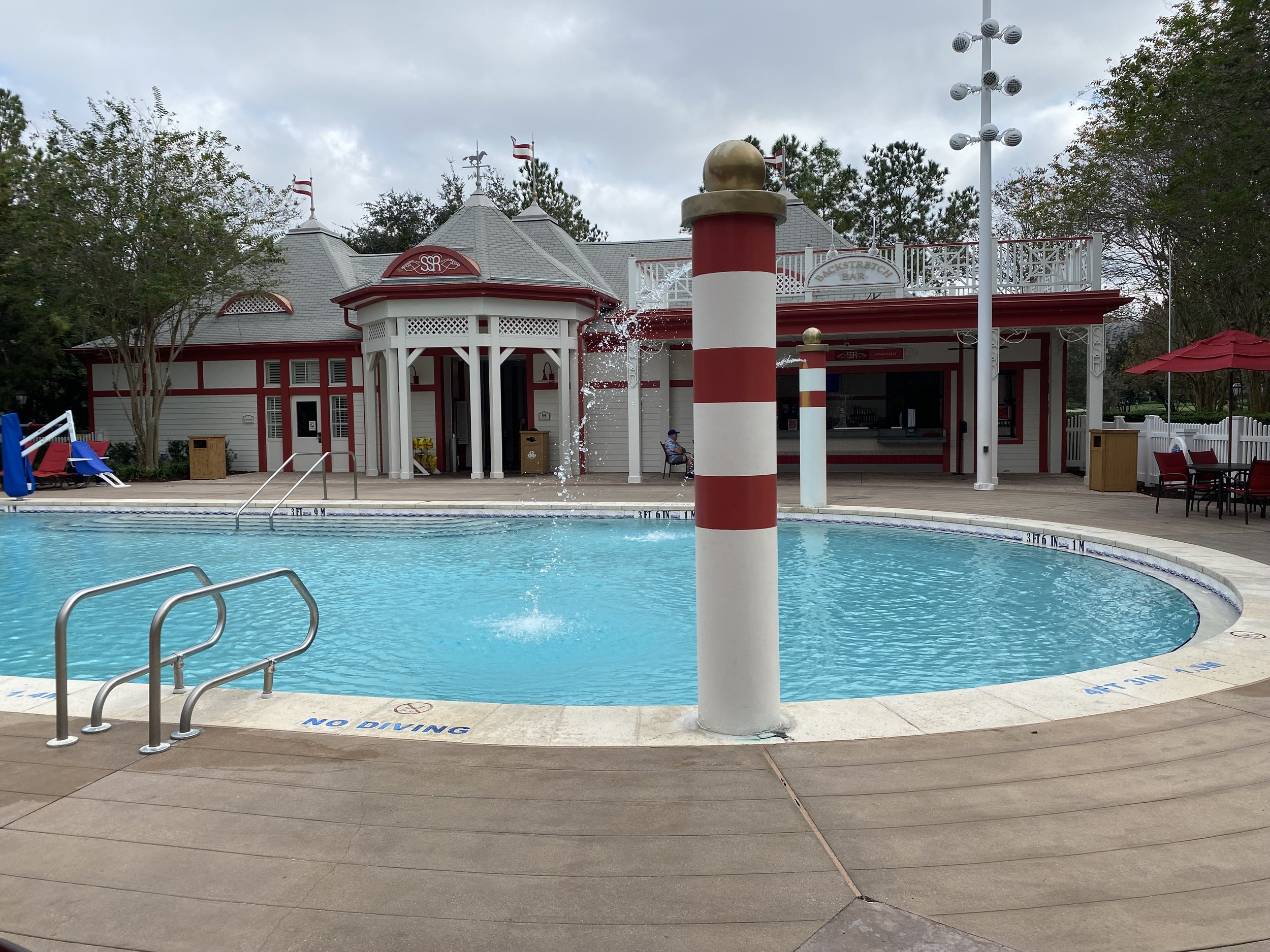 Empty swimming pool with a water fountain and a striped pole in the center, surrounded by a deck with chairs and umbrellas, building in the background with a sign reading 'Backstretcher Bar'.