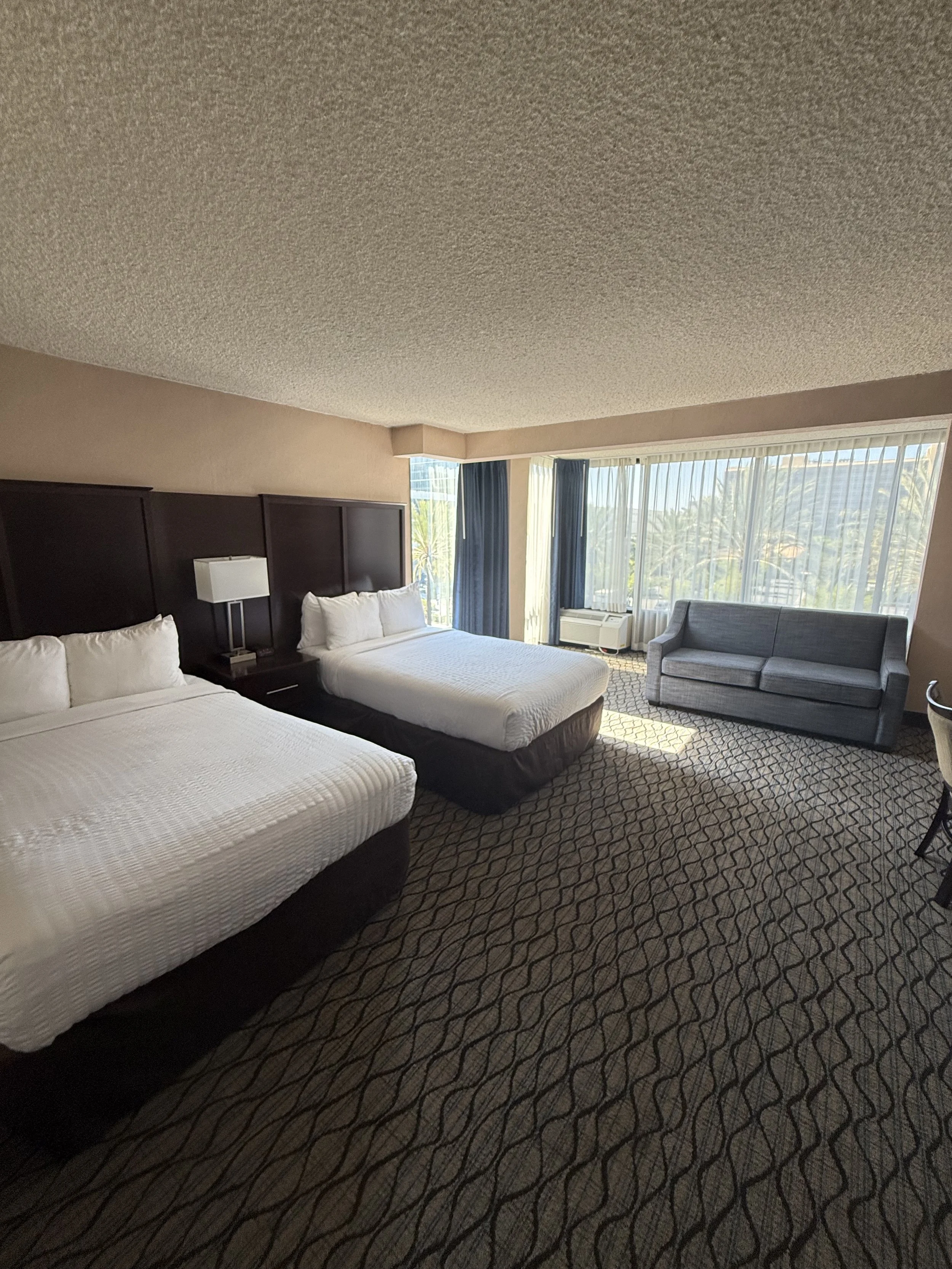 A hotel room with two beds, a gray sofa, a large window with curtains, and a patterned carpet.