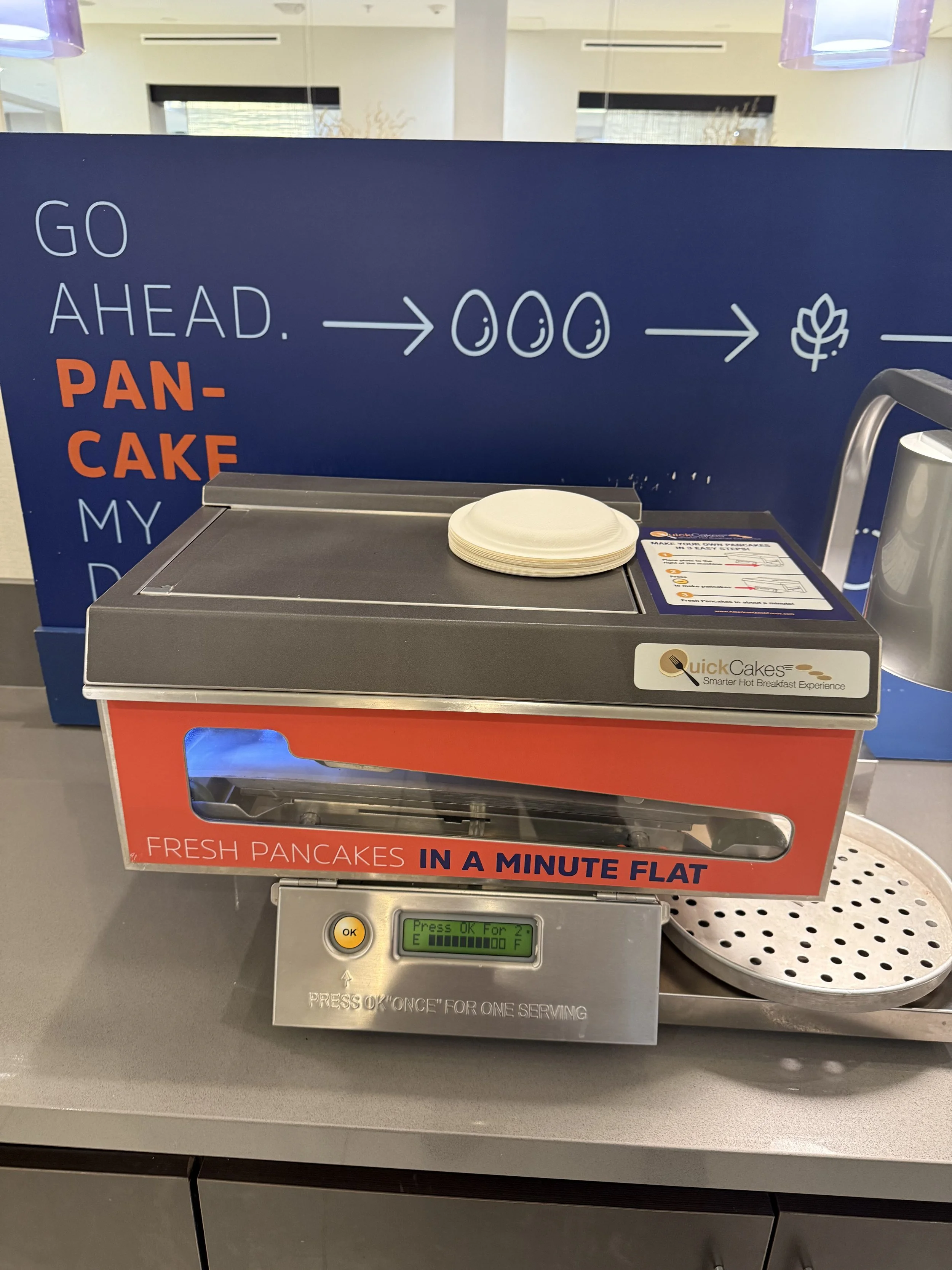 A pancake machine with a sign behind it that says "Go Ahead. Pancake My Day." The machine offers fresh pancakes in a minute and has a digital display and a button to press for a serving.